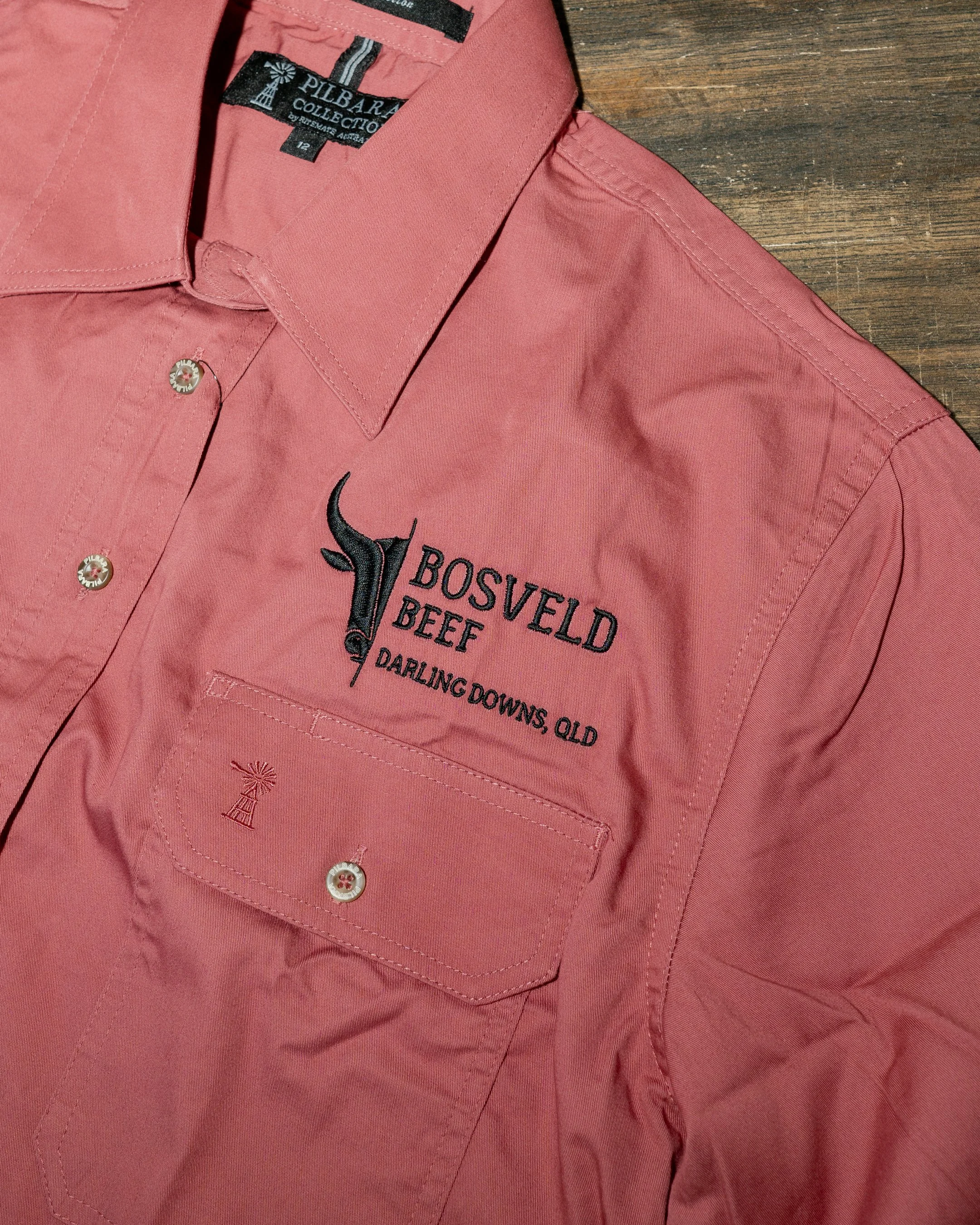 Close-up of a pink button-up shirt with embroidered text and logo, showing part of the collar and chest pocket, placed on a wooden surface.