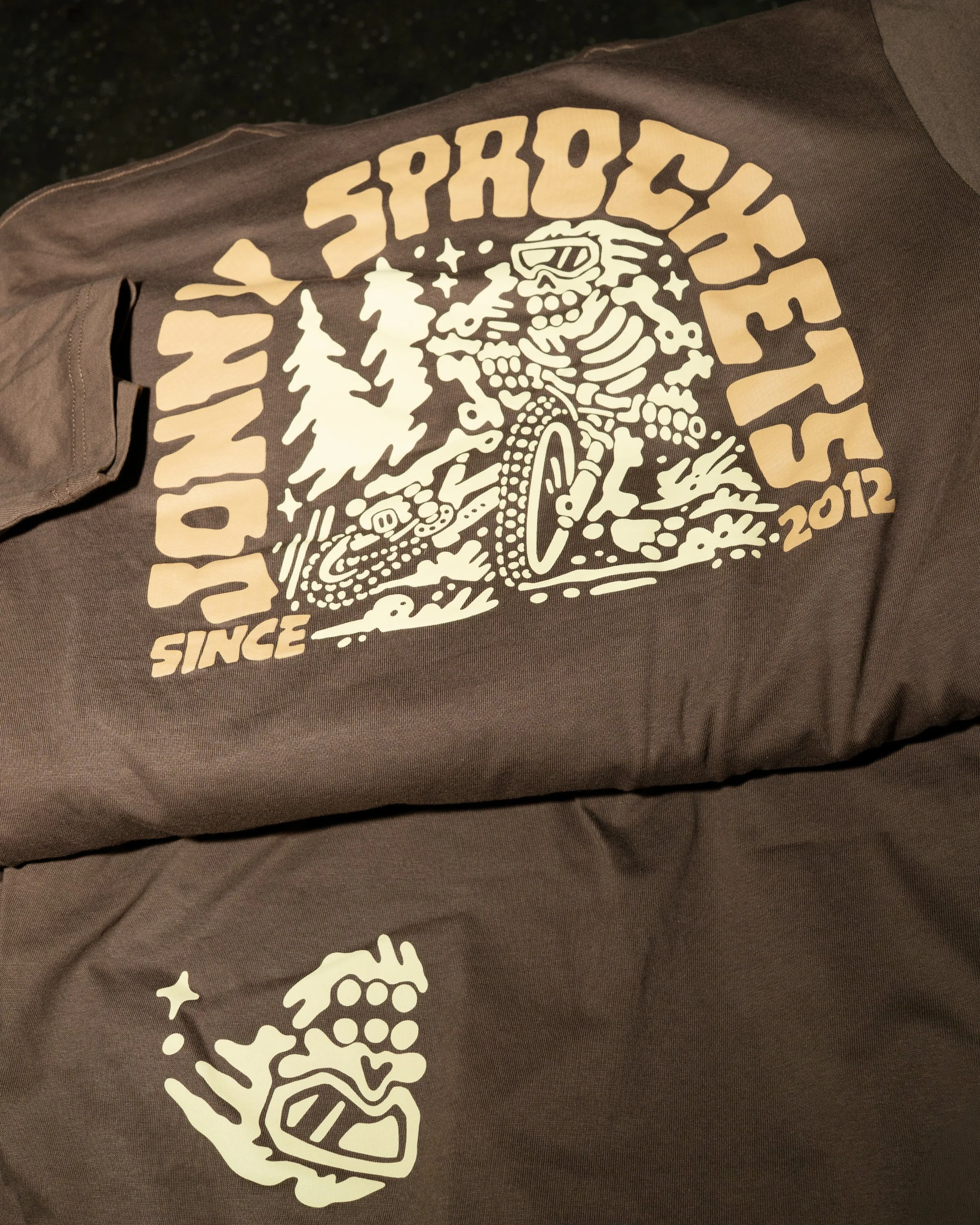 Brown t-shirt with a graphic design featuring a skeleton wearing sunglasses, surrounded by trees and stars, with the text "SINCE 2012" and other stylized lettering.