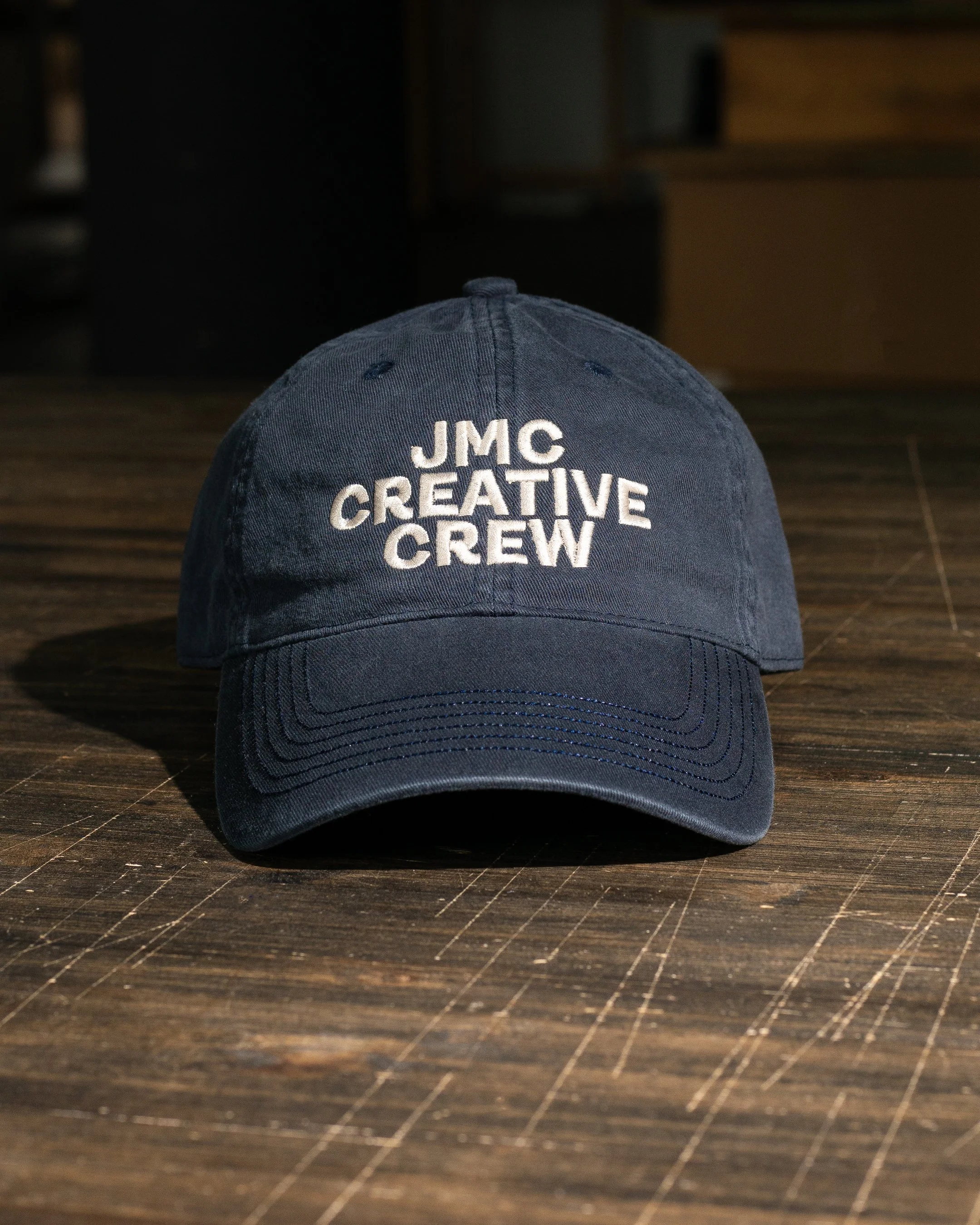 Close-up of a navy blue baseball cap with white embroidered text that reads 'JMC CREATIVE CREW' on the front, placed on a dark wooden surface.