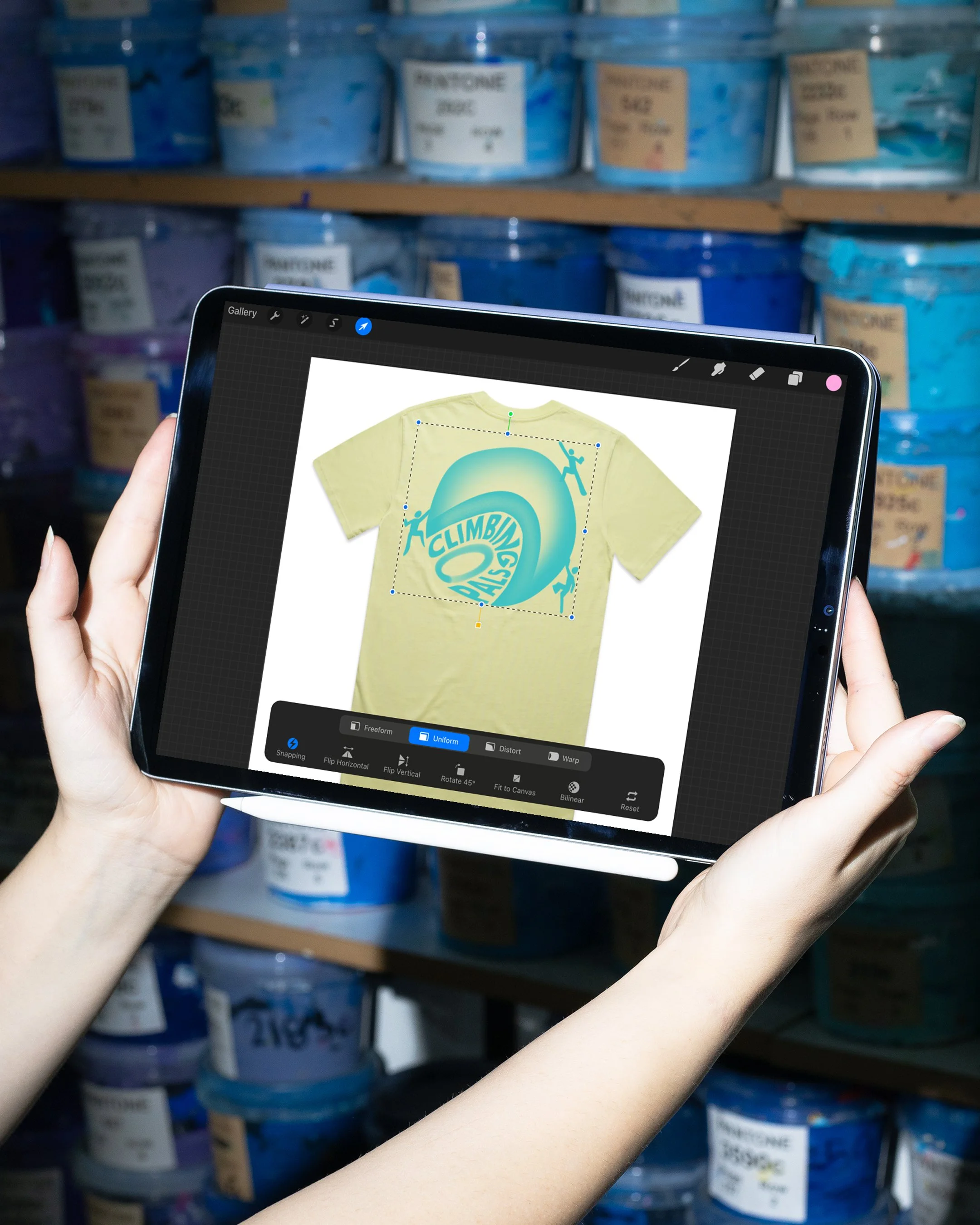Person holding a tablet with a t-shirt design of a yellow t-shirt with a blue and green wave and climbing figures on it, in a screen printing shop with shelves of blue and purple ink tubs in the background.
