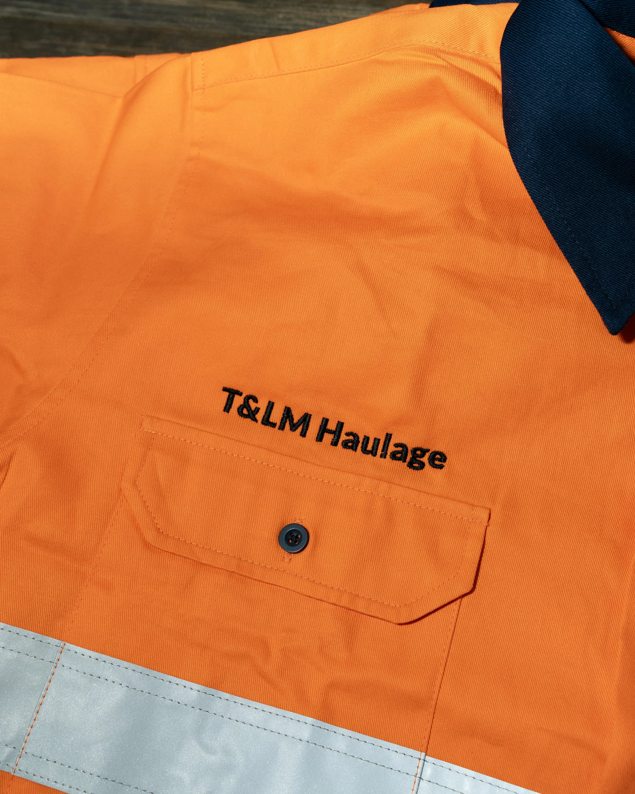 Close-up of an orange safety vest with a black button, black text reading 'T&L M H', and a dark blue collar, resting on a gray surface.