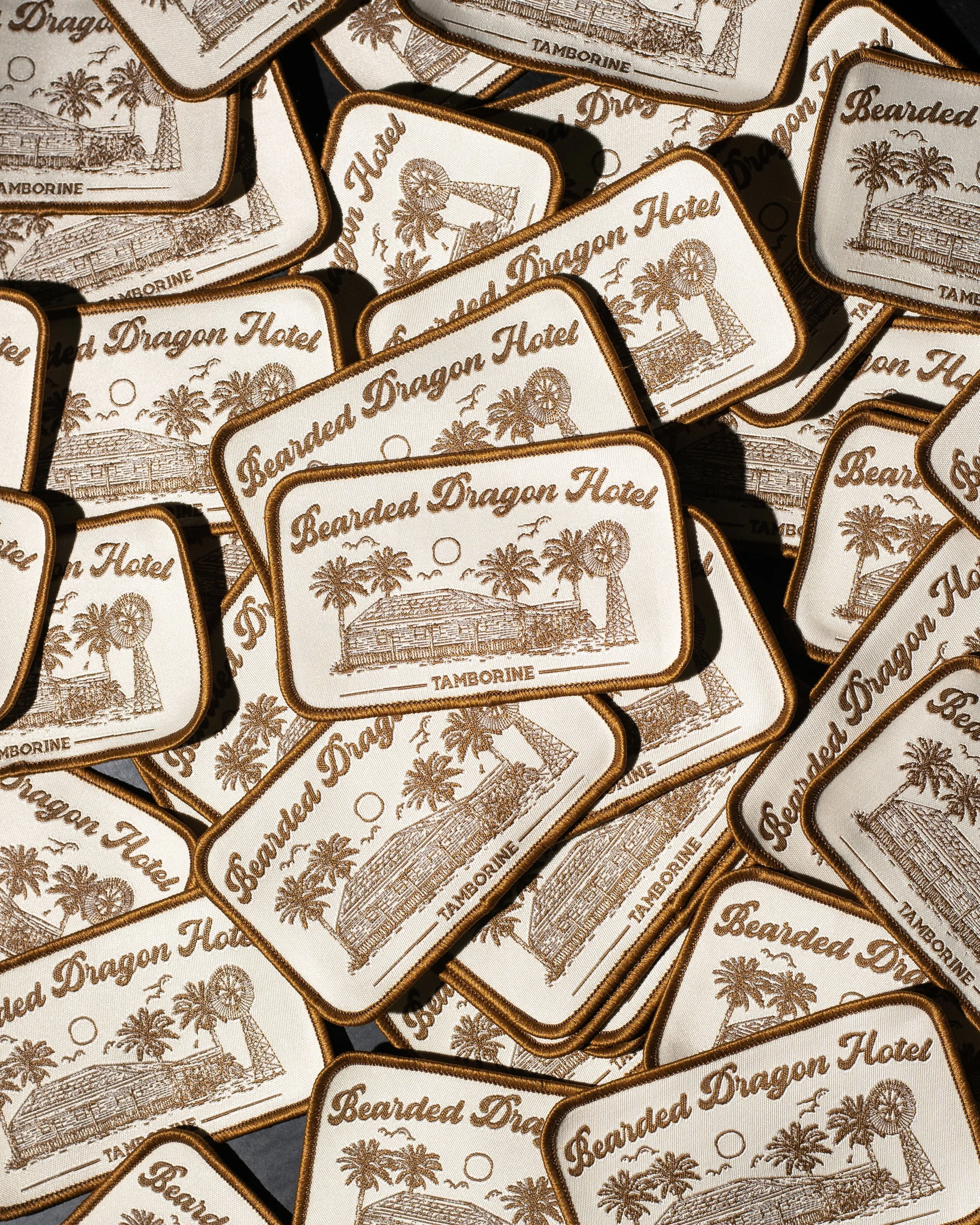 Multiple embroidered patches with a beige and brown design featuring a building, palm trees, a windmill, and the text 'Bearded Dragon Hotel' and 'TAMBOURINE'.