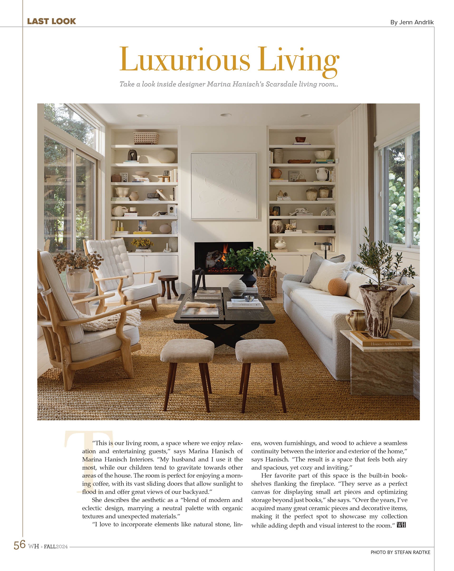 A cozy living room designed by Marina Hanisch, featuring built-in bookshelves flanking a fireplace, contemporary furniture, and various decorative objects. The space is illuminated by large windows, offering a view of greenery outside. Neutral tones 