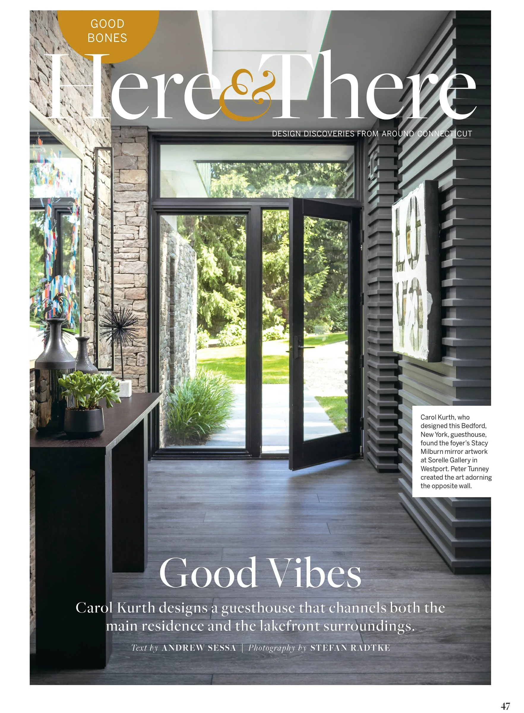 Interior view of a modern guesthouse foyer with large glass doors and stone walls, designed by Carol Kurth. The space includes decorative items on a table and artwork by Peter Tunney on the wall.