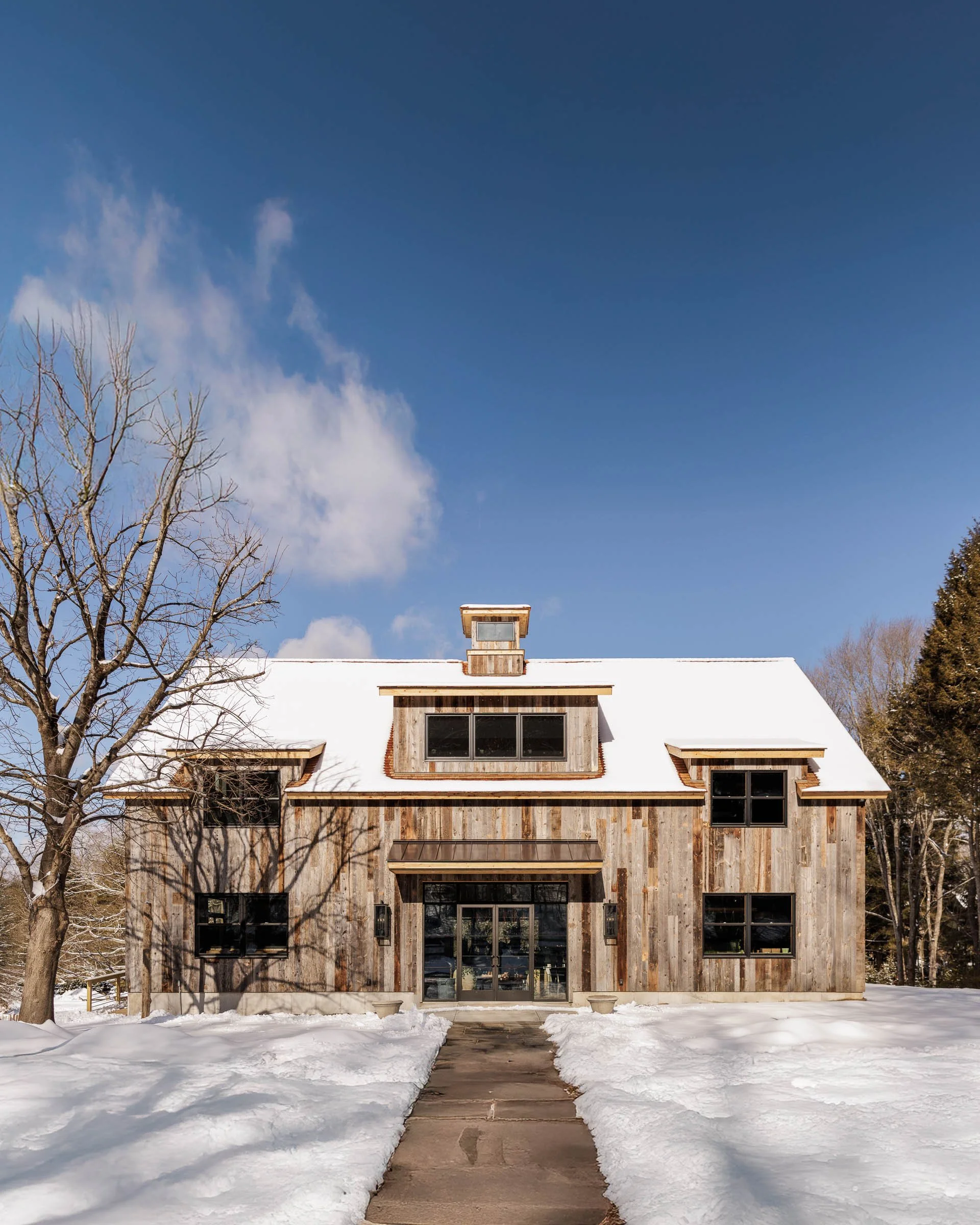 Wooden house with snow-covered roof and path, bare tree on left, against blue sky with clouds.