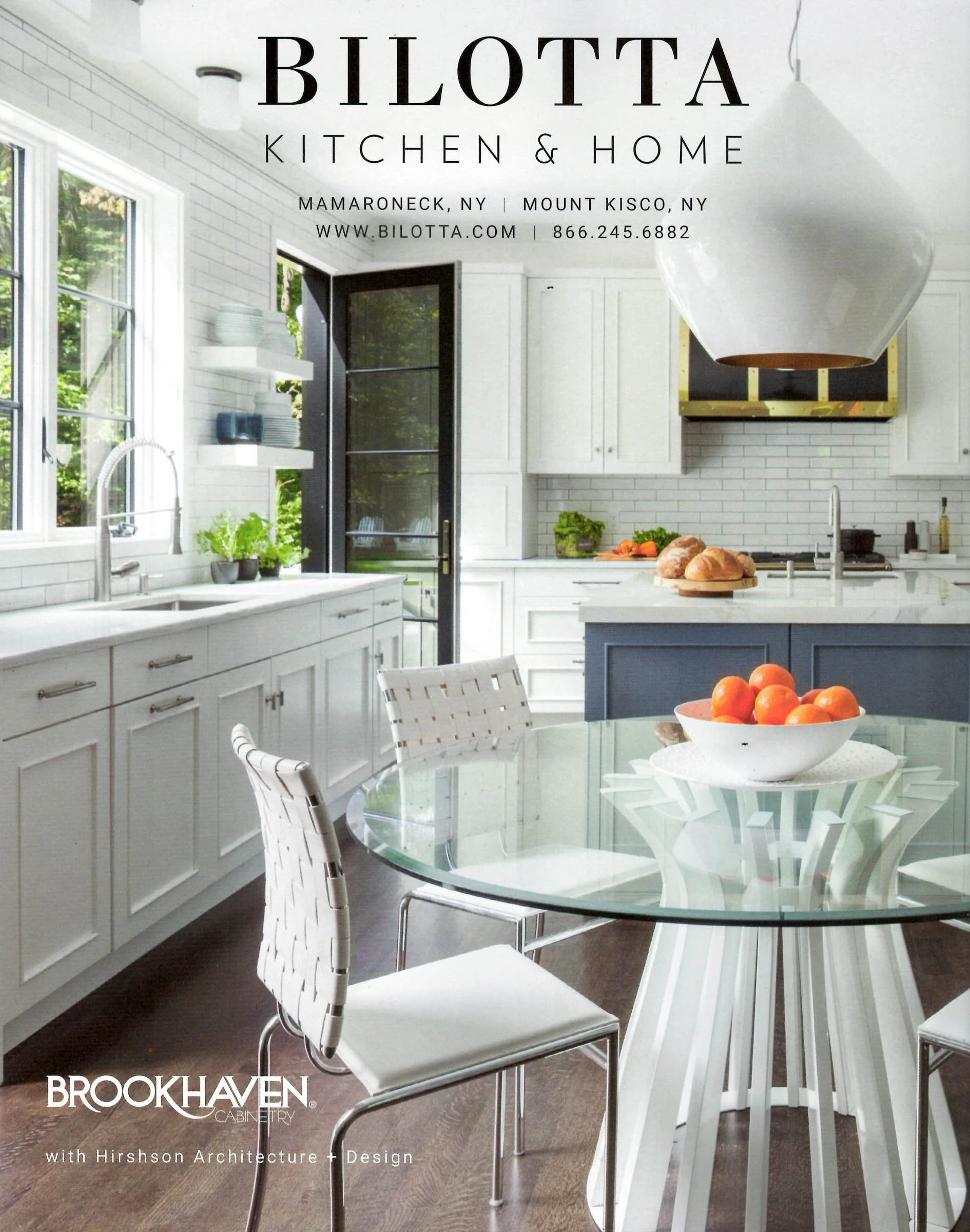 Modern kitchen with white cabinets, glass table, white chairs, fruit bowl with oranges, stainless steel appliances, and large pendant light.