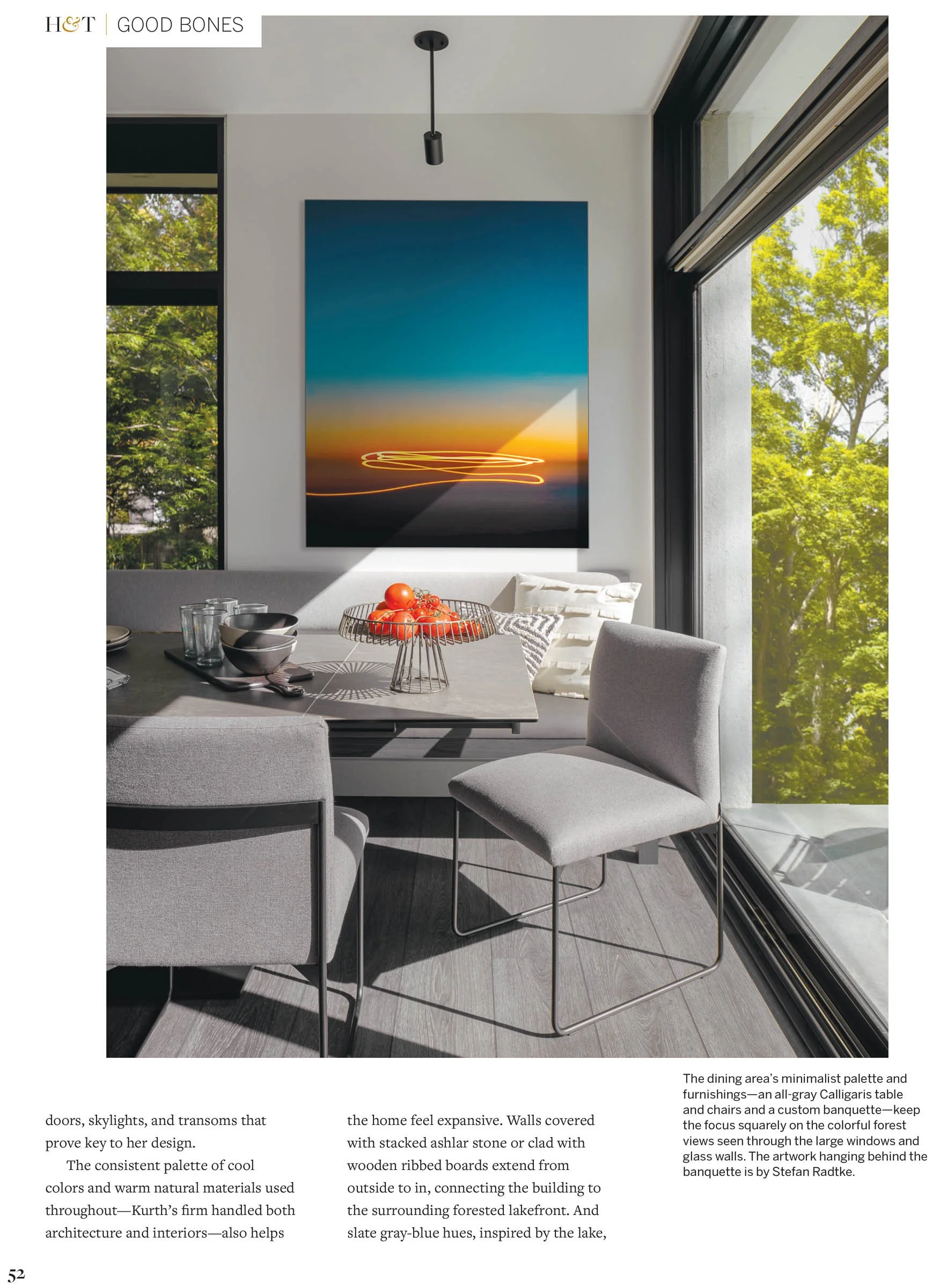 Modern dining area with a minimalist design, featuring a large abstract artwork on the wall, gray chairs, a gray table, and a glass bowl of tomatoes. Large windows reveal green trees outside.