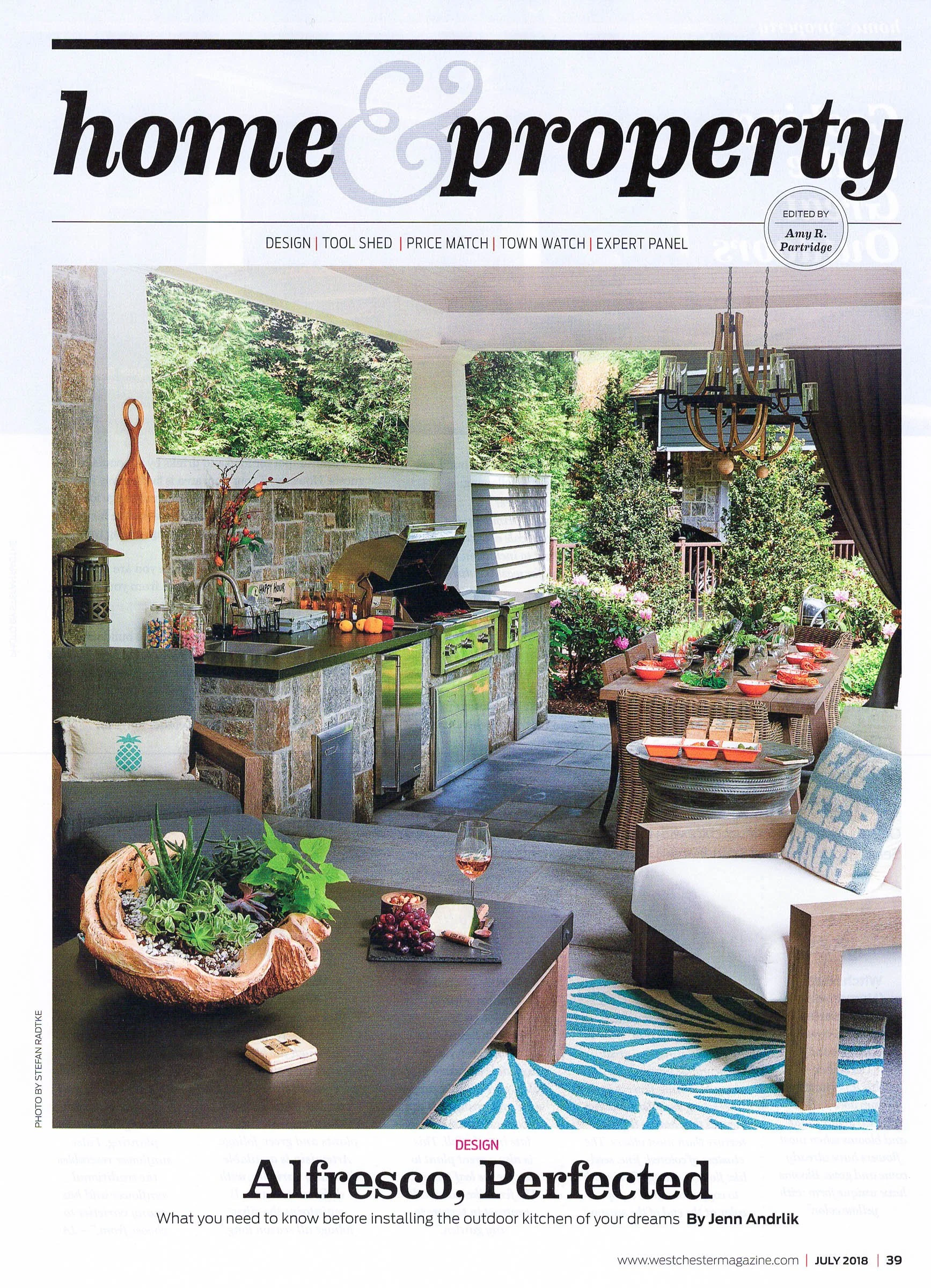 Outdoor kitchen with stone countertops, grill, wooden dining table set, and lounge chairs. Lush greenery surrounds the area. "Home & Property" magazine cover with title "Alfresco, Perfected."