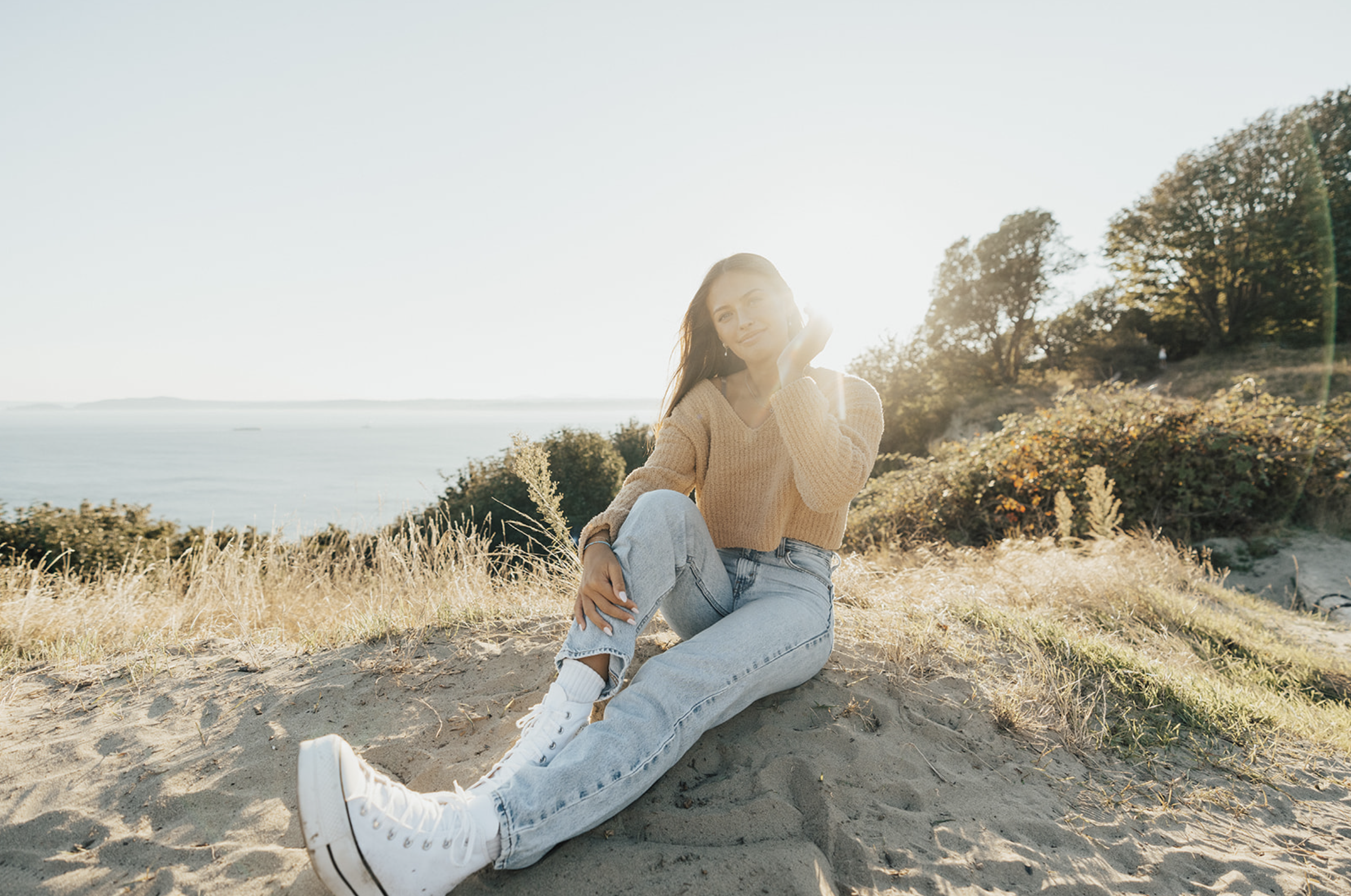 Young woman sitting on sandy hilltop near water with trees in the background, wearing a beige sweater and white sneakers, illuminated by sunlight.