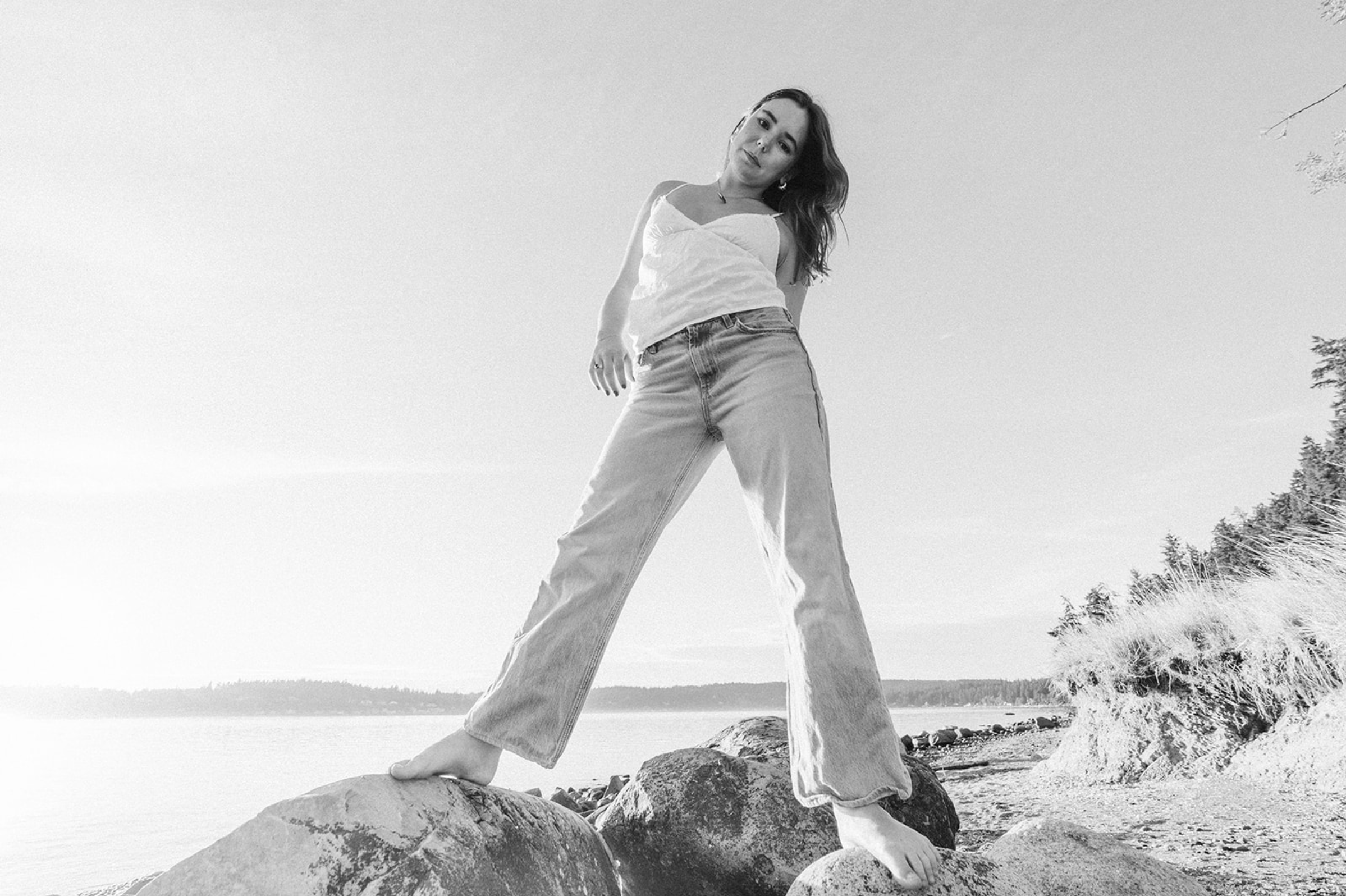A young woman standing on large rocks at a lakeshore, looking down at the camera, wearing a white tank top and wide-leg jeans, with trees and water in the background.