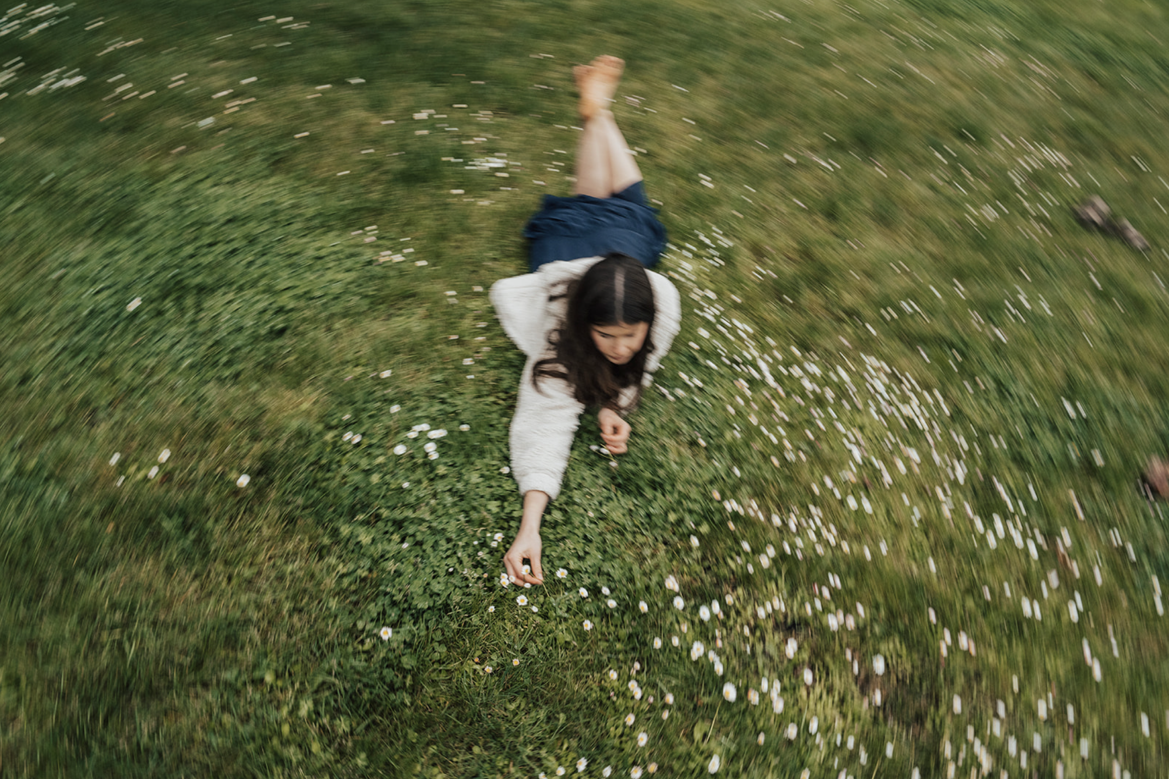 A girl lying on the grass in a field of small white flowers, reaching out to pick one, motion blur creates a swirling effect around her.