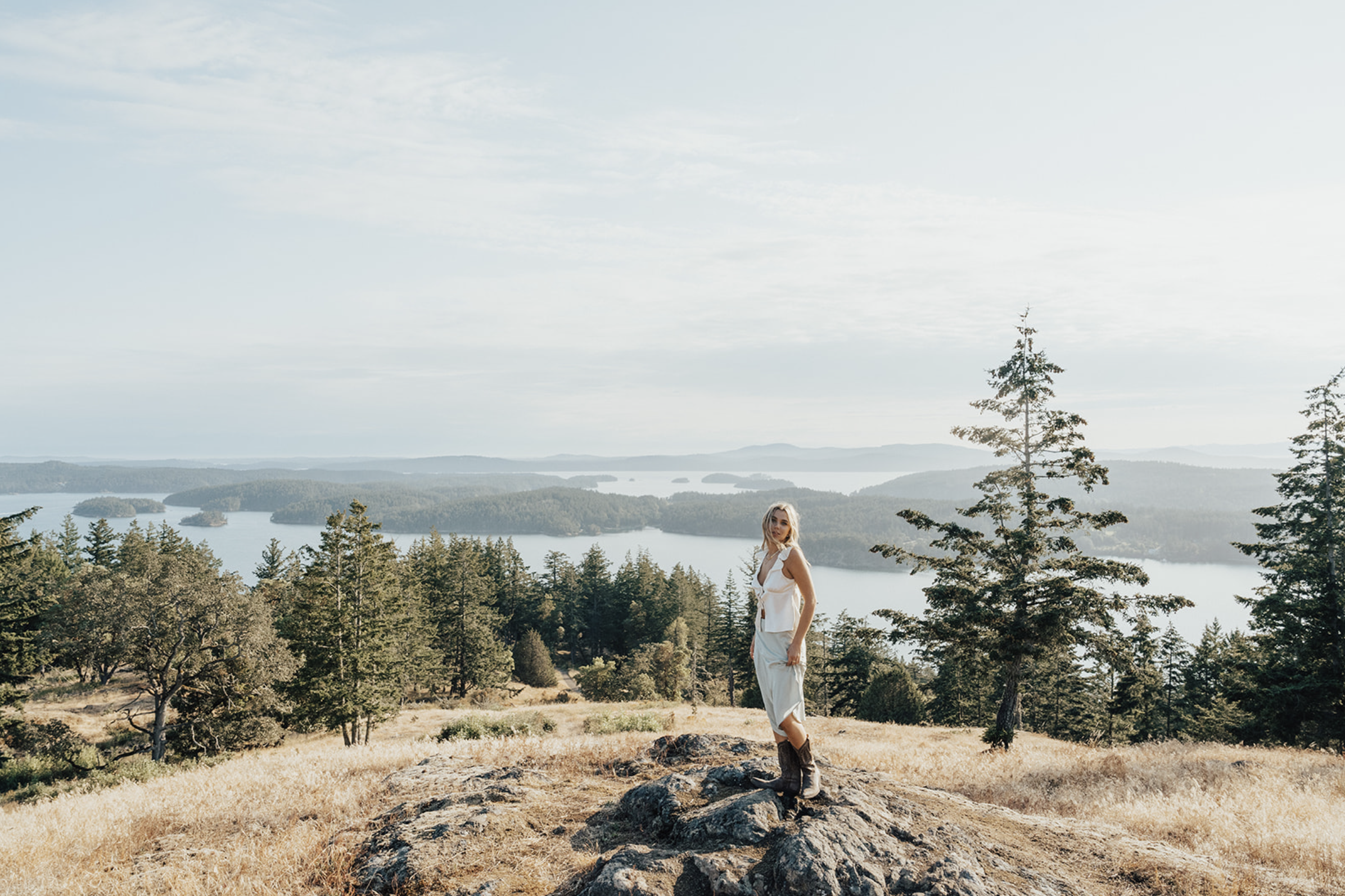 A woman standing on a rock in a wooded area overlooking a large body of water with islands and distant hills under a partly cloudy sky.
