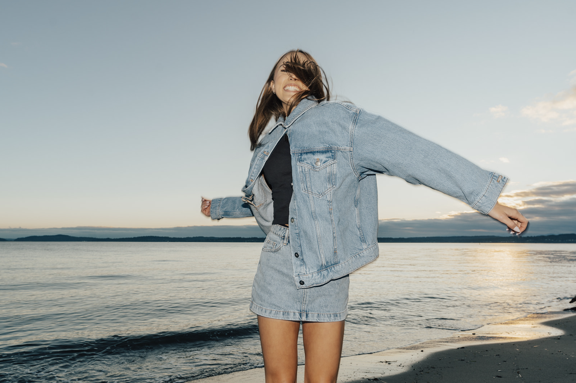 A woman smiling and joyfully spinning on a beach during sunset, wearing a denim jacket and skirt.