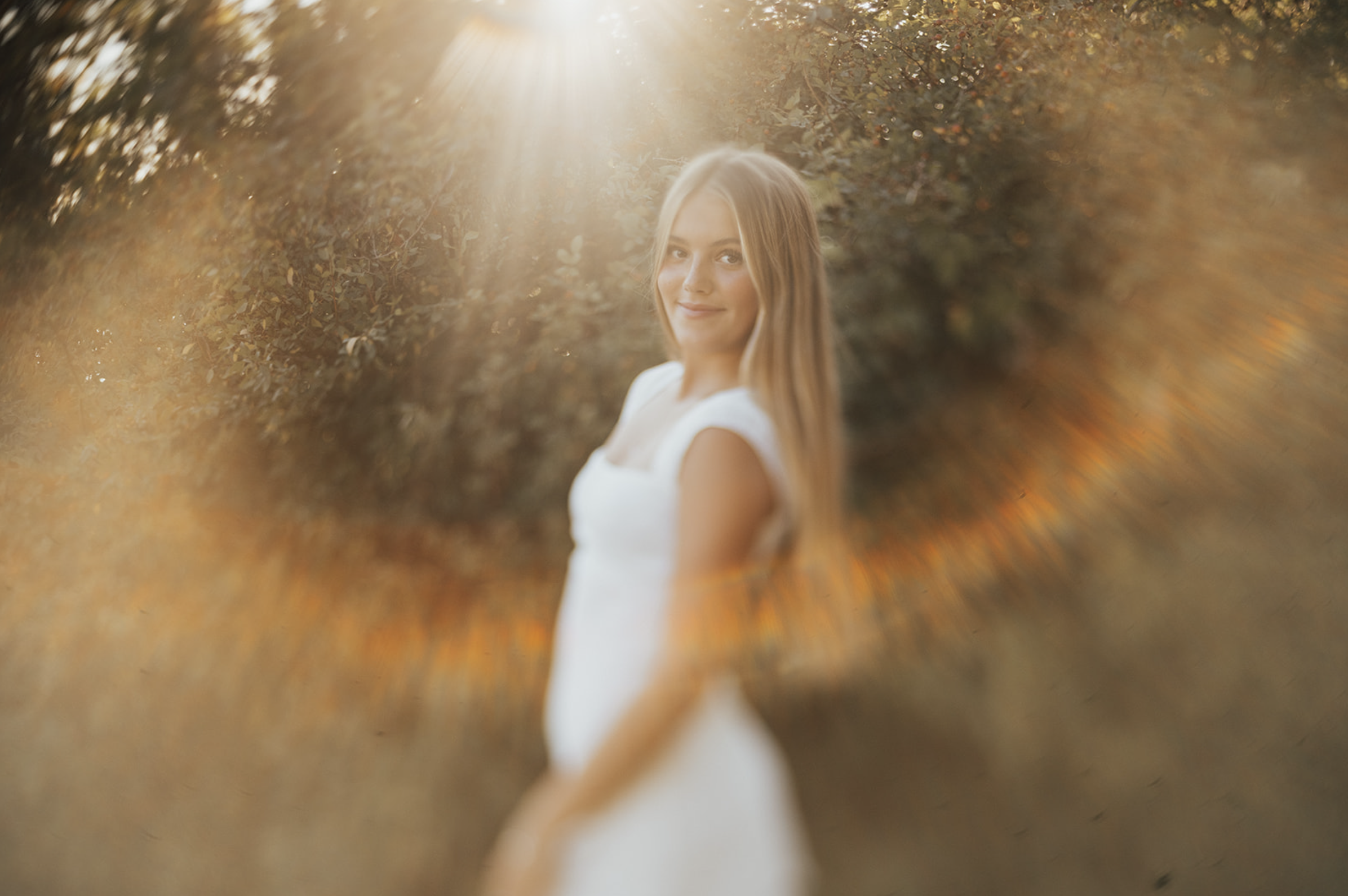 A young woman with long blonde hair standing outdoors during sunset, smiling and looking at the camera, with a blurred background of trees and warm light.