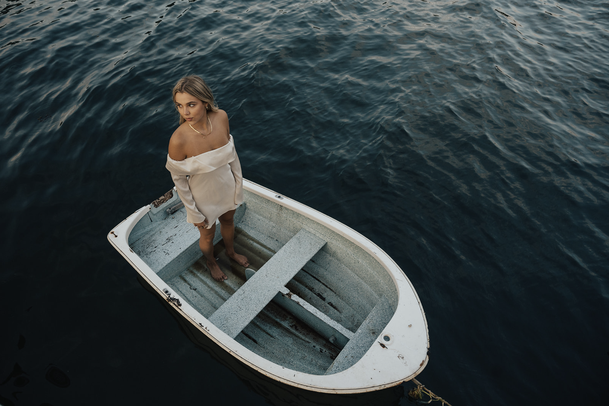 A woman with blonde hair standing barefoot in a small, empty boat on dark water, looking upwards to her left, wearing an off-shoulder white dress.