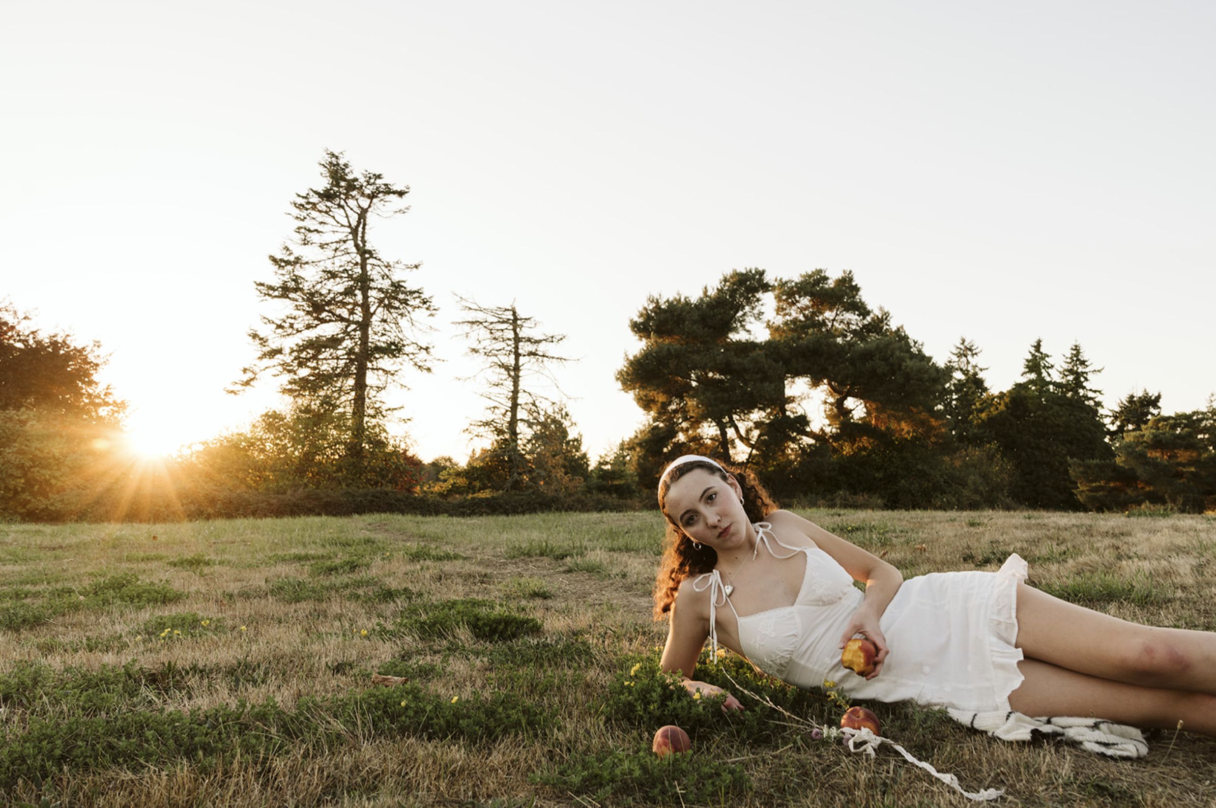 A young woman in a white dress lying on the grass in a field at sunset, holding an apple and surrounded by a few more apples.