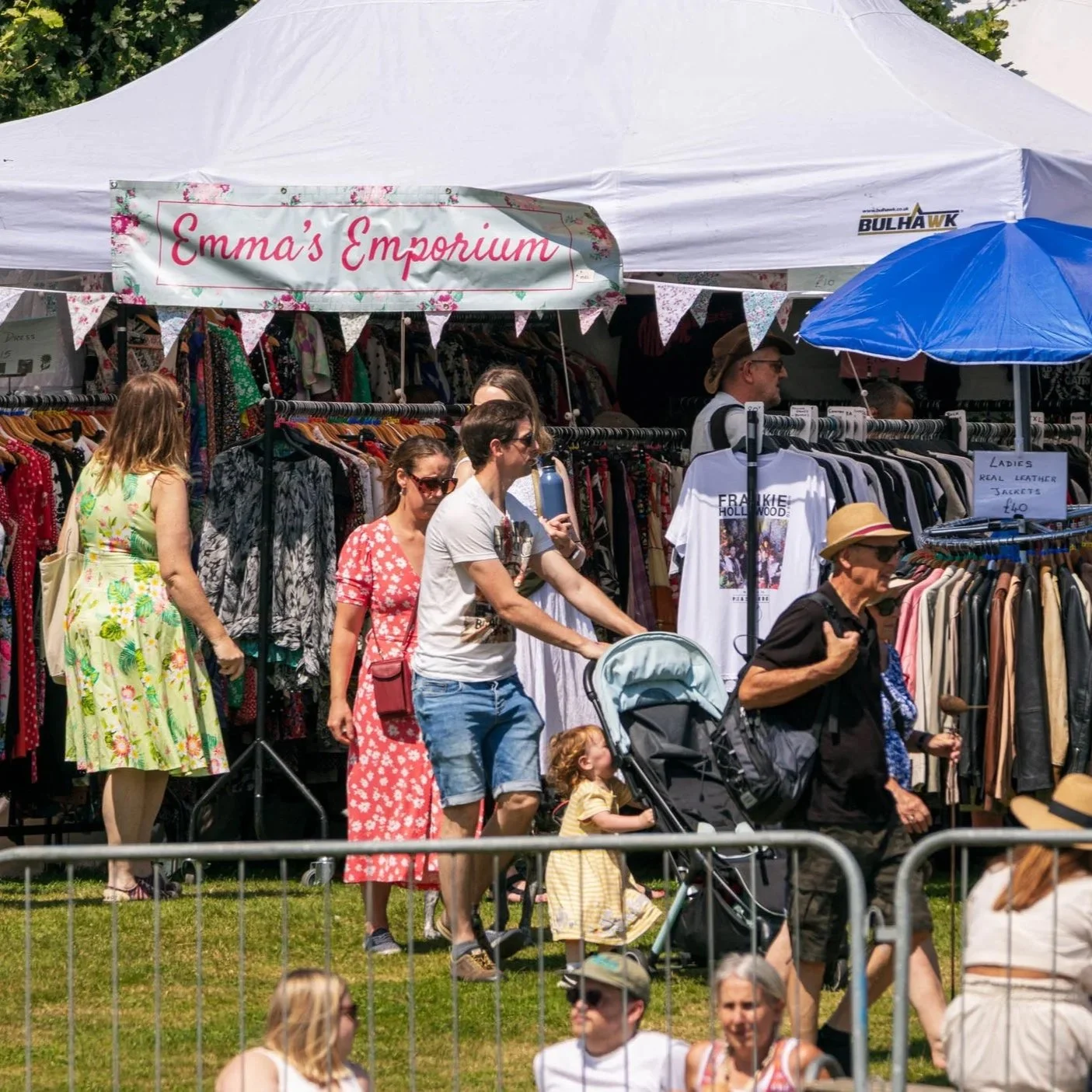 Public walking around the festival on grass, past Emma's Emporium clothing stand
