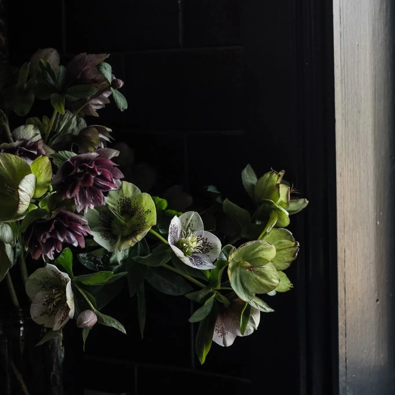 A bouquet of flowers with light hitting them, placed in front of a dark tiled wall and a wooden surface on the right side.