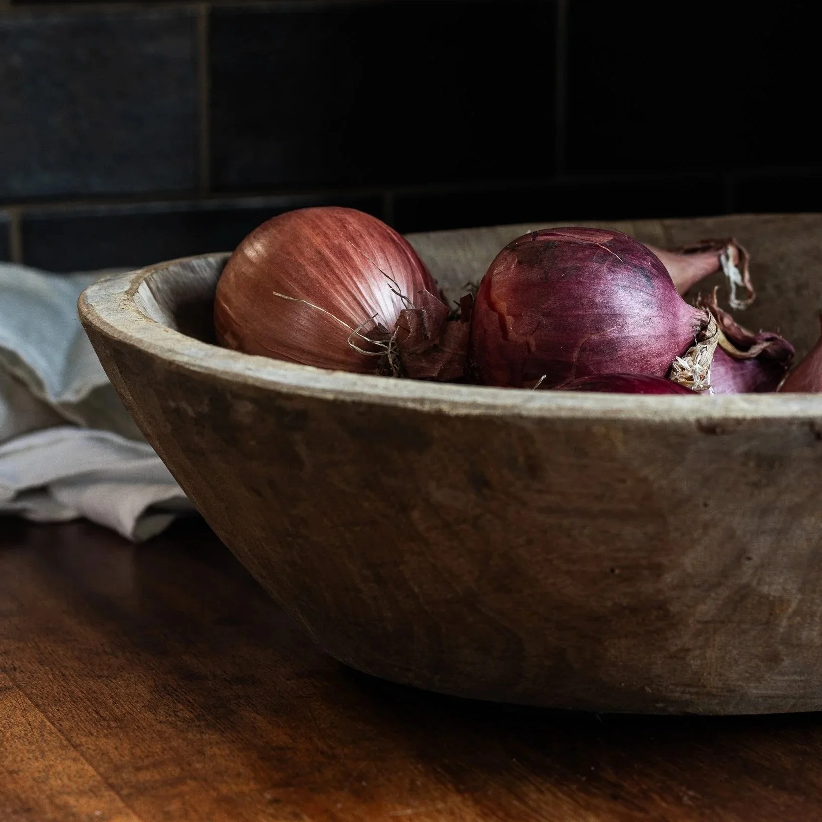 A wooden bowl containing red onions, placed on a wooden surface with a white cloth nearby, against a dark background.