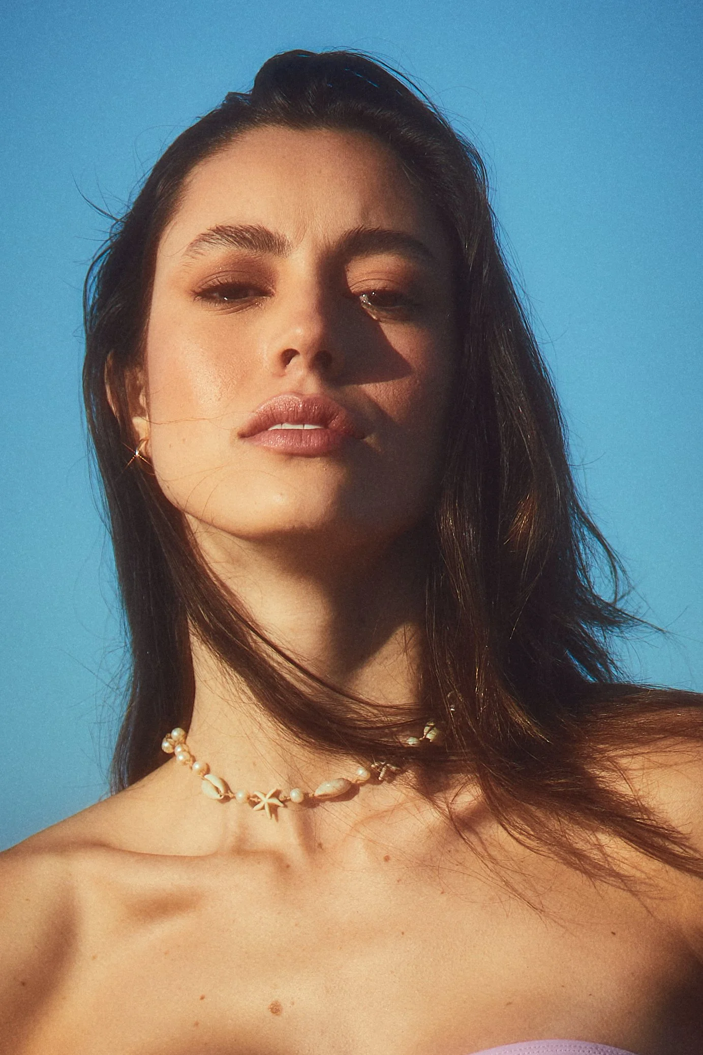 Close-up of a woman's face with dark hair, wearing a pearl necklace and earrings, against a blue background.