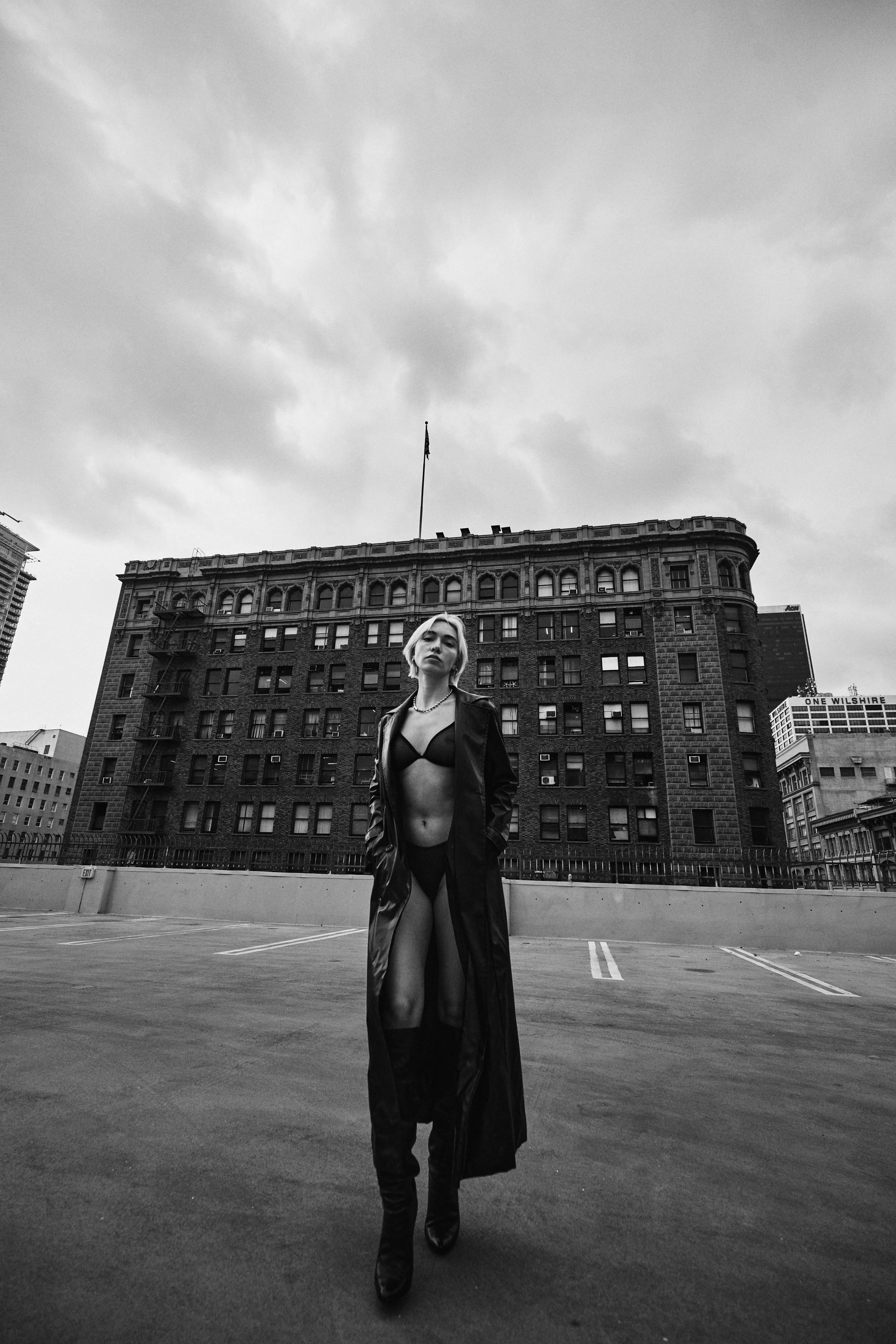 A woman walking confidently in an empty rooftop parking lot. She is dressed in a black bra, long open coat, and black boots. The background features a tall, old brick building with a flag on top and a cloudy sky.