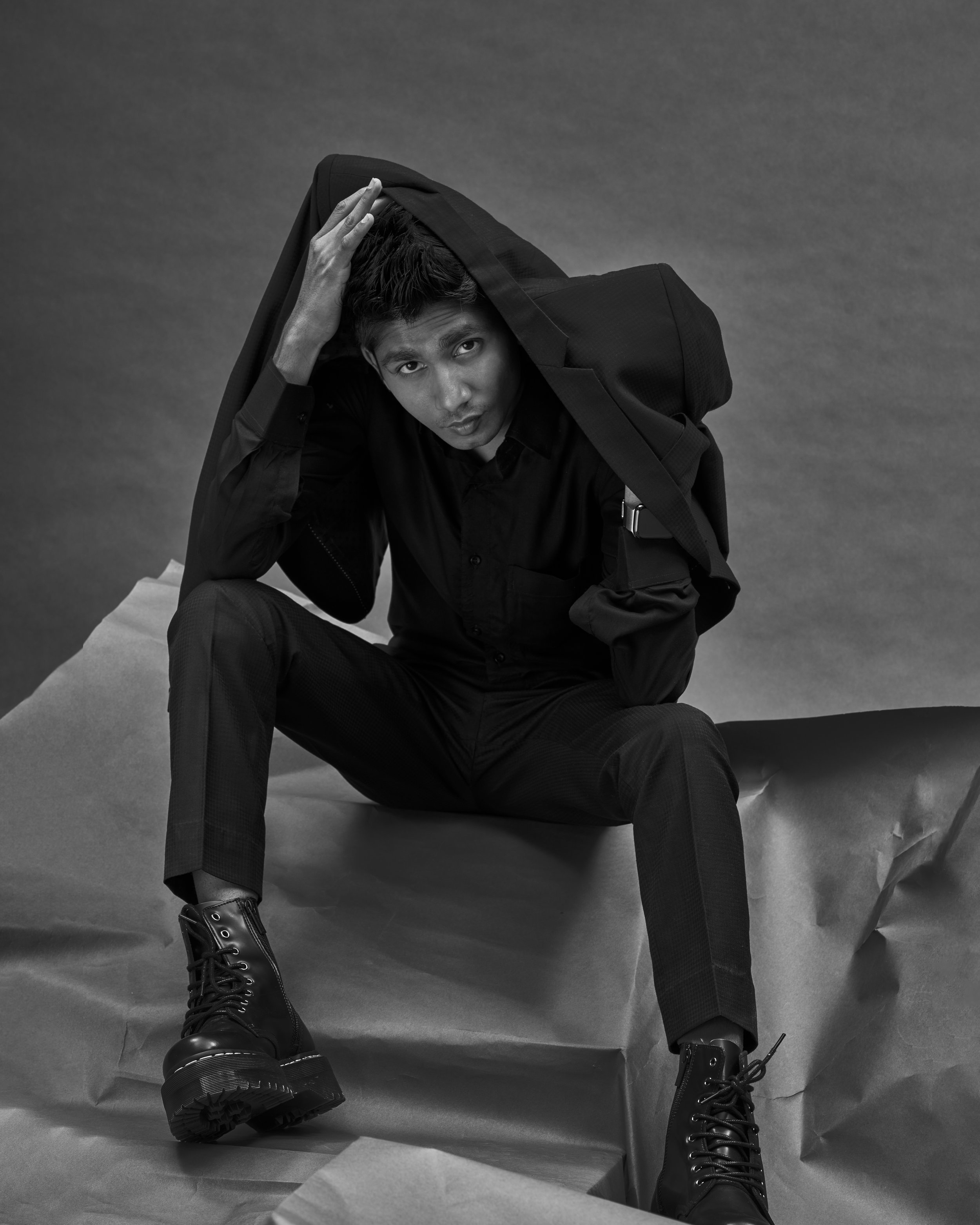 A young man dressed in black, sitting on the floor, with a black backpack on his shoulder, fixing his hair with his left hand, wearing black combat boots, in front of a plain, textured backdrop.