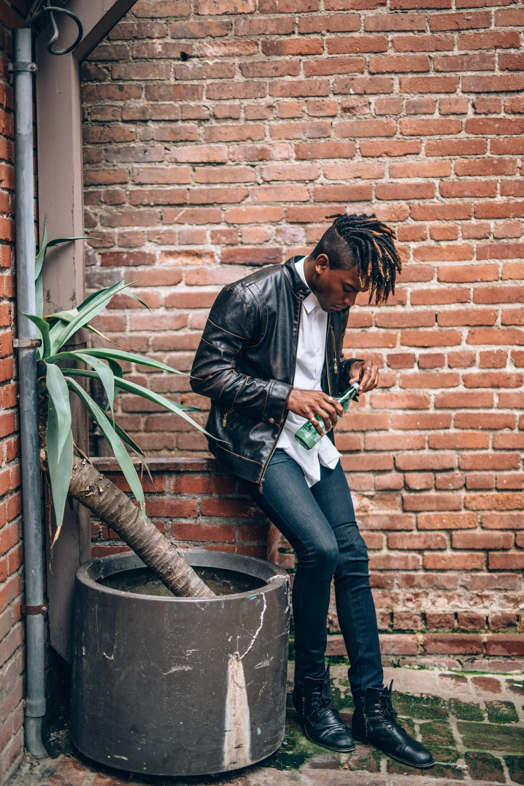 A man with dreadlocks wearing a black leather jacket, white shirt, jeans, and black boots standing against a brick wall, looking at a bottle in his hand.