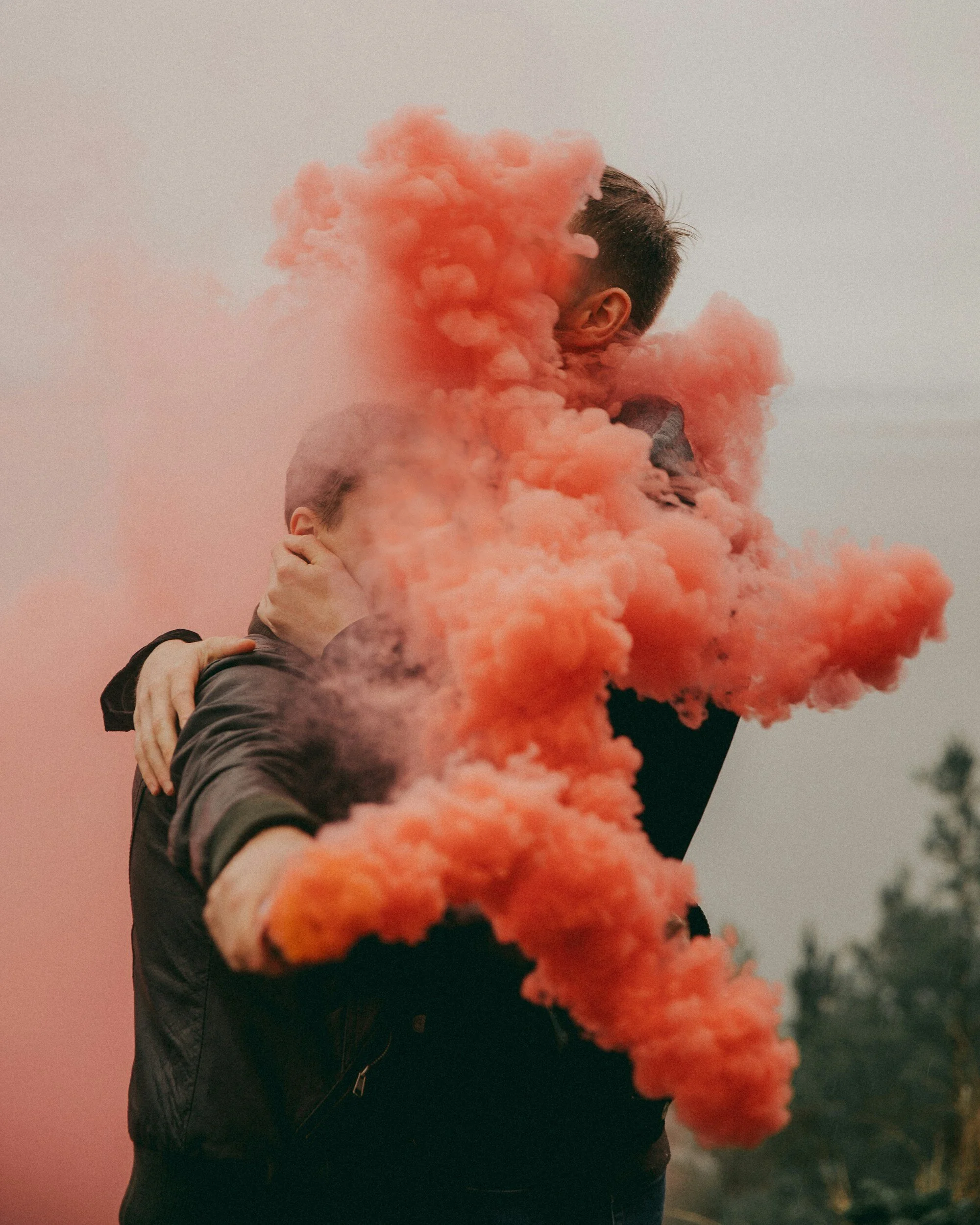 Two people embracing while holding a pink smoke bomb, which is billowing thick pink smoke between them.