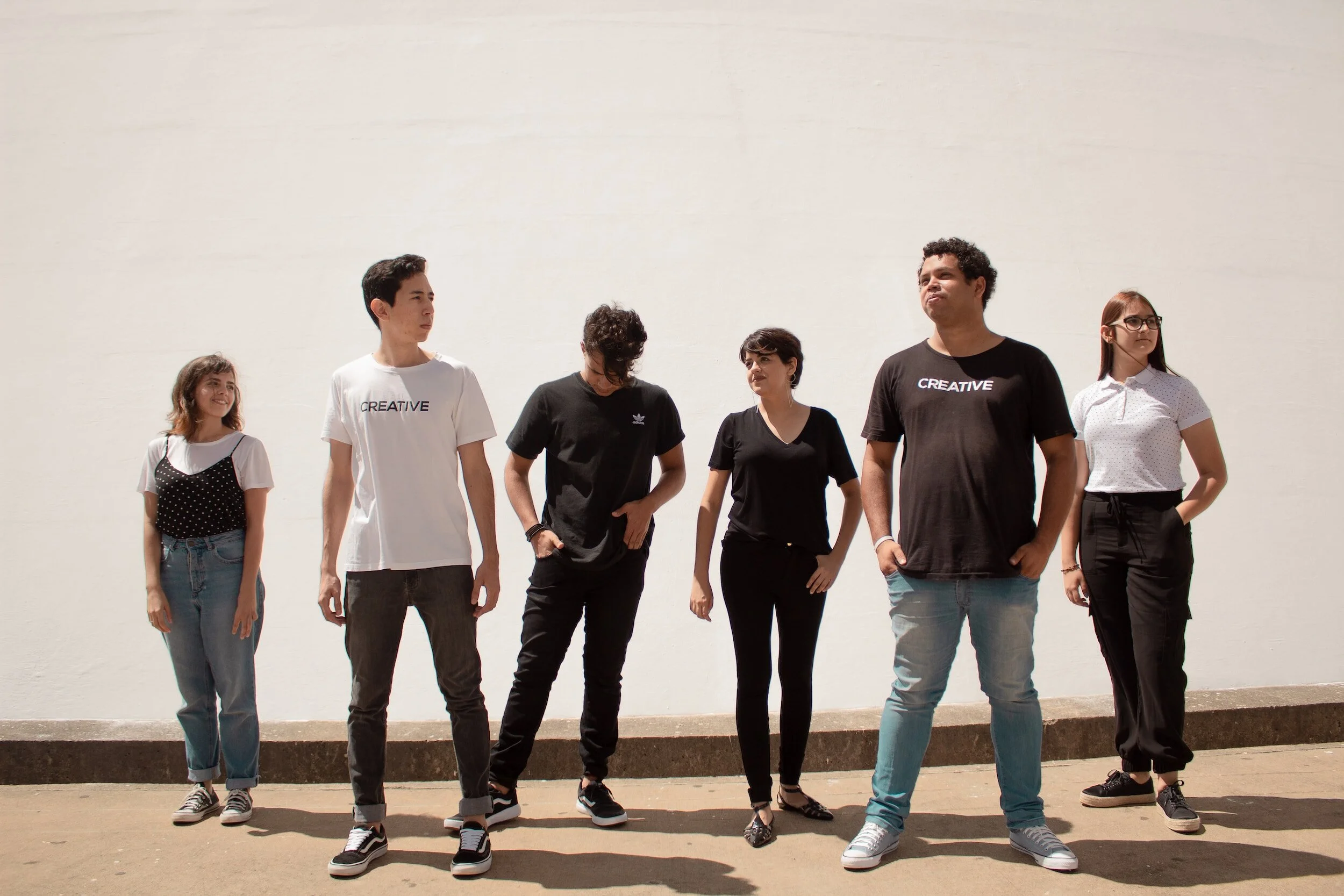 Group of six young adults standing outdoors against a plain white wall, some wearing casual clothes, with two saying 'CREATIVE' on their T-shirts.