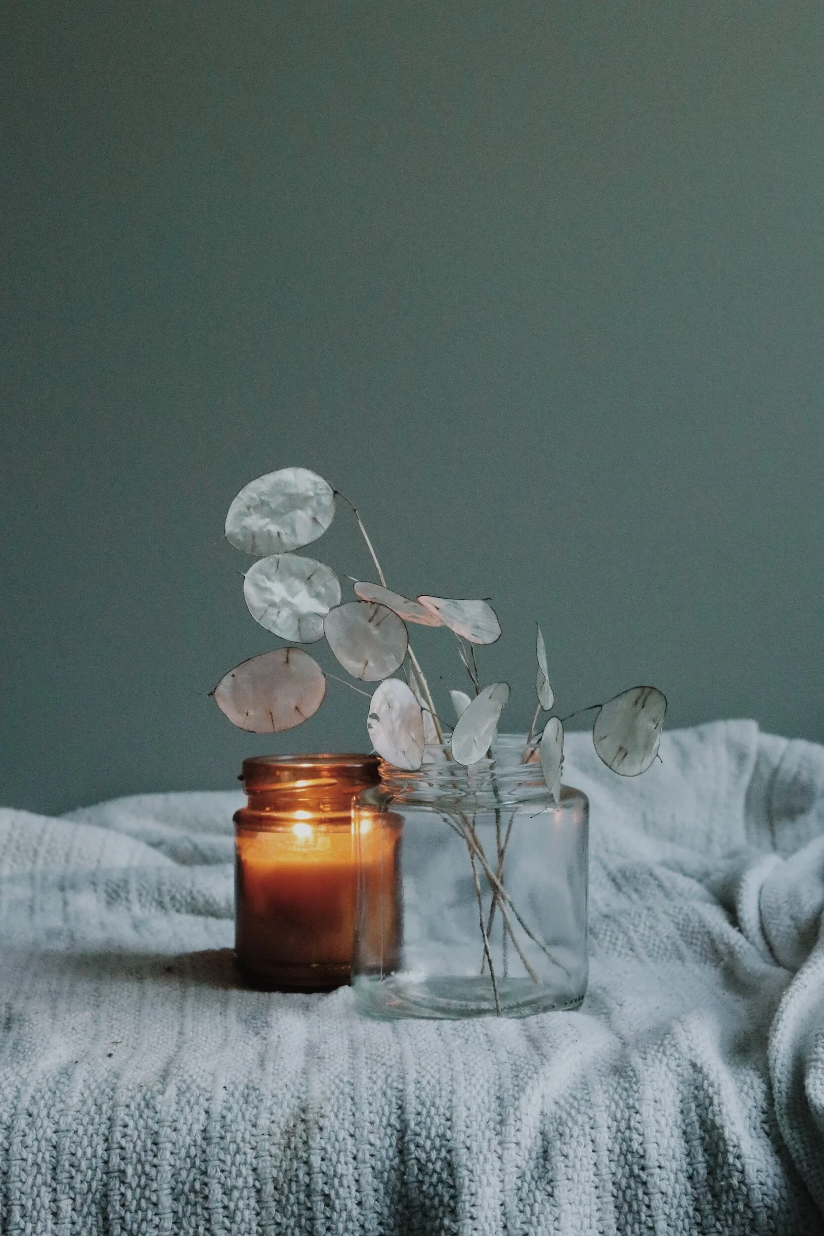 A glass jar with a dried eucalyptus arrangement beside a lit amber candle on a textured white fabric surface with a plain muted green background.