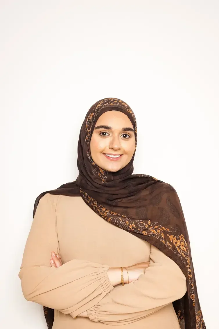 A young woman wearing a brown hijab with orange and beige patterns, beige long-sleeved top, standing with arms crossed, smiling at the camera against a plain white background.