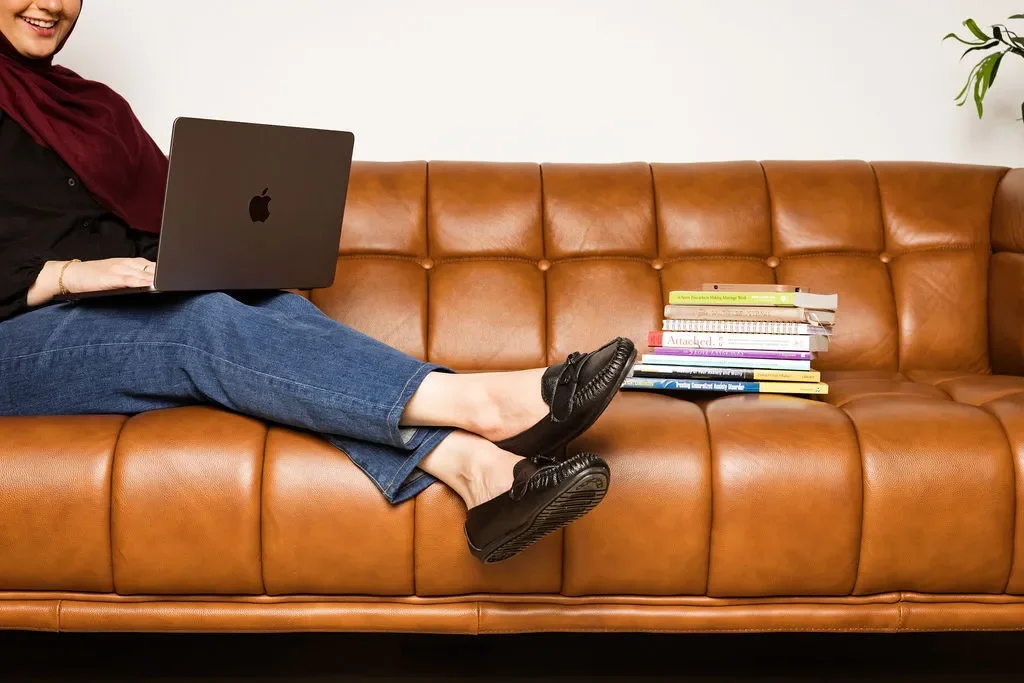 A woman sitting on a tan leather couch with a laptop on her lap and a stack of books beside her.