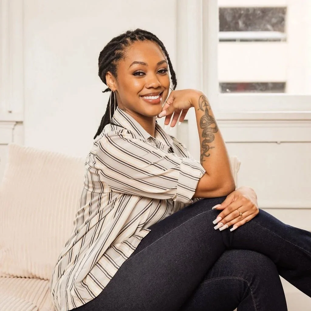 A woman with dreadlocks, smiling and resting her face on her hand, wearing a white sleeveless top, with tattoos on her arms, sitting on a sofa with a white textured cover.