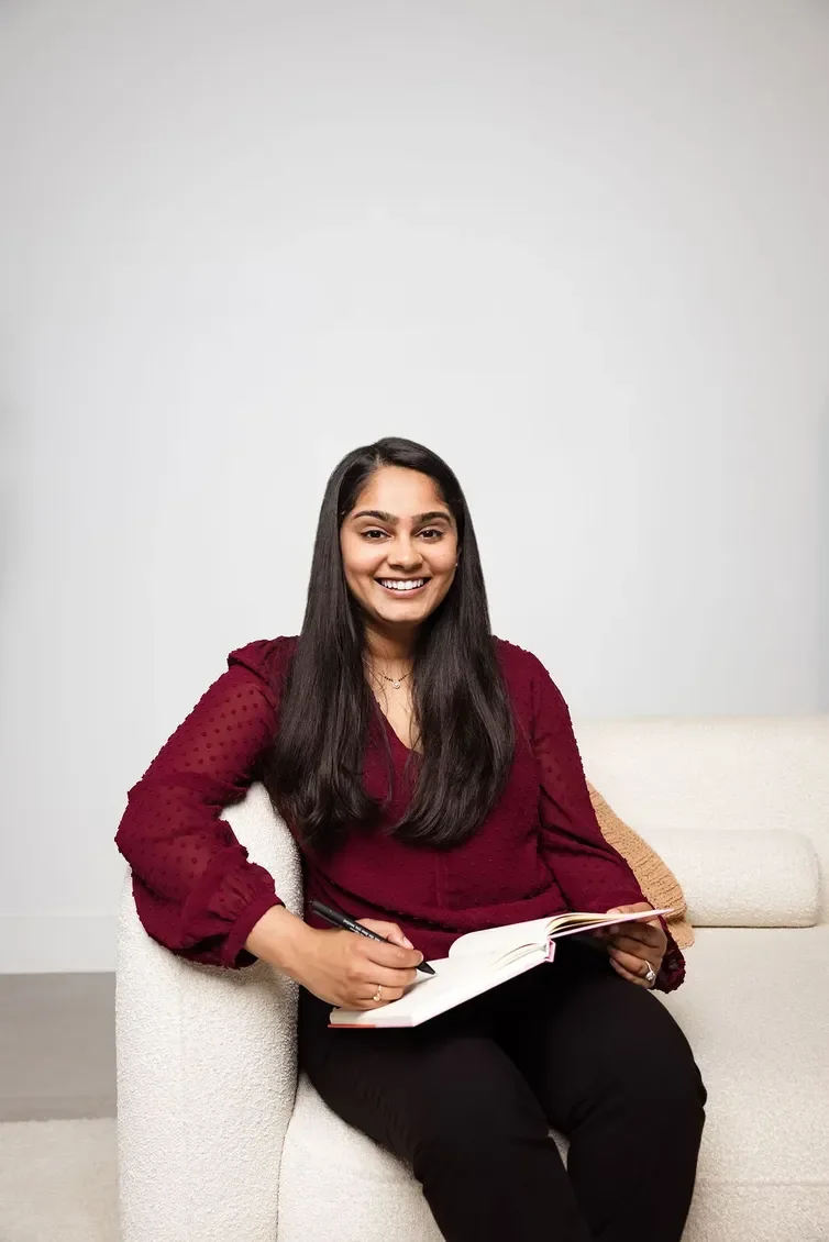 A woman with long dark hair sitting on a white couch, smiling, holding a notebook and pen, wearing a burgundy blouse and black pants.