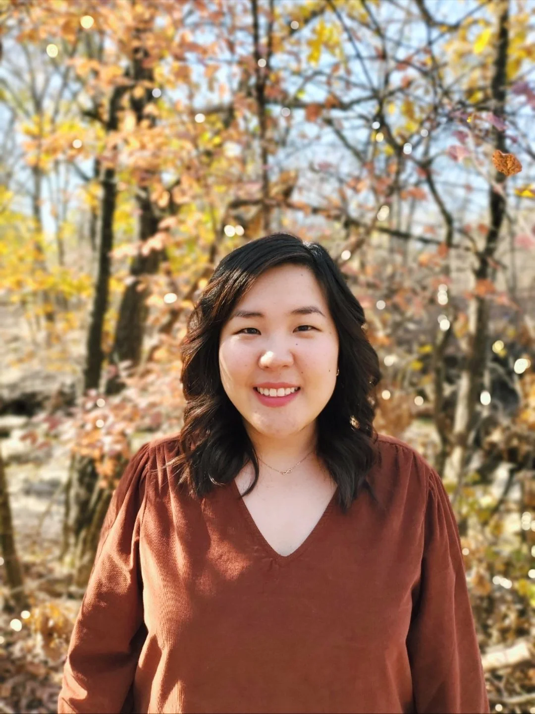 A woman with dark hair smiling outdoors in autumn, standing in front of trees with colorful fall leaves.