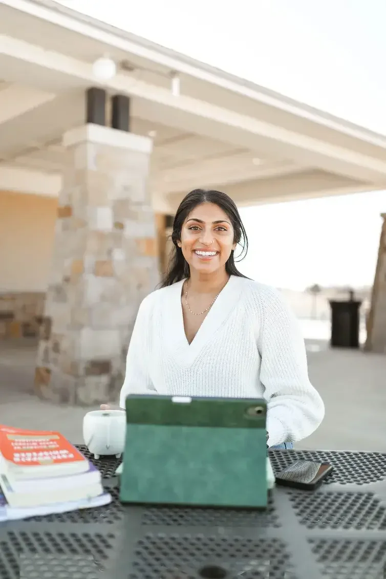 A woman with dark hair and a white sweater smiling while sitting at an outdoor table with a tablet, a cup, and books.