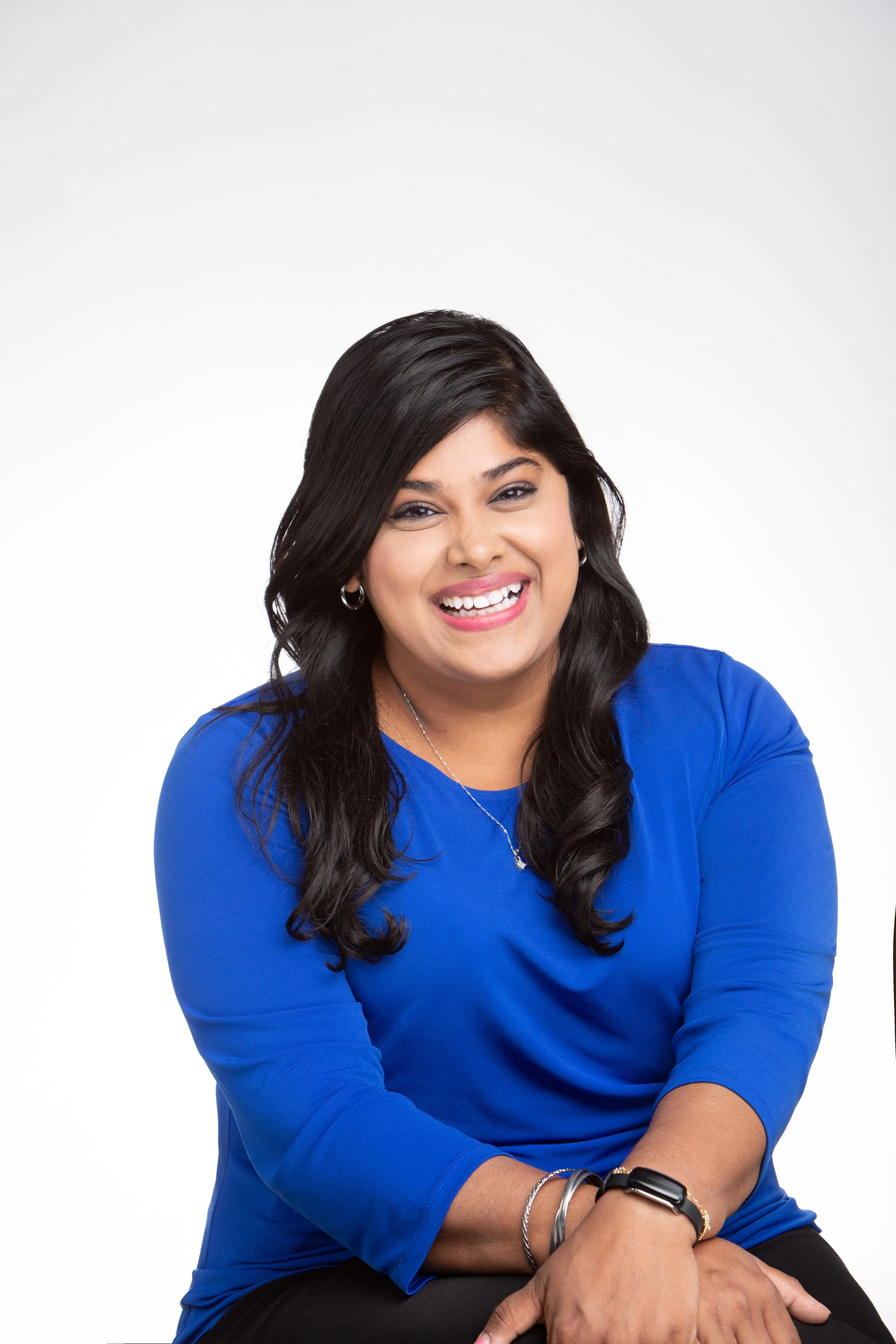 A smiling woman with long dark hair wearing a blue shirt, silver jewelry, and a black watch, posing against a plain white background.