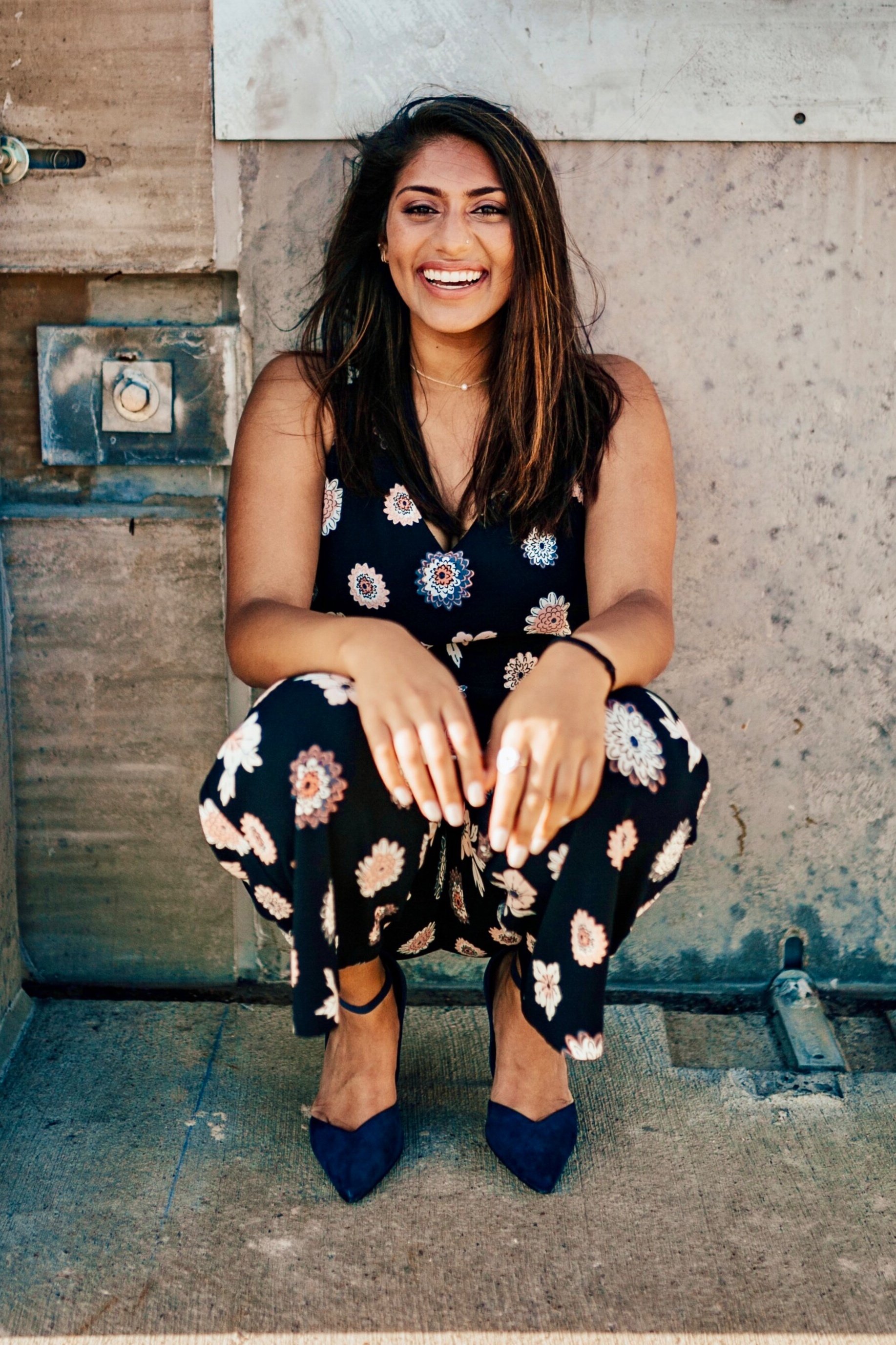 A woman with dark hair and a bright smile, wearing a navy floral jumpsuit and matching navy heels, is crouching and sitting on a concrete floor against a weathered wall.