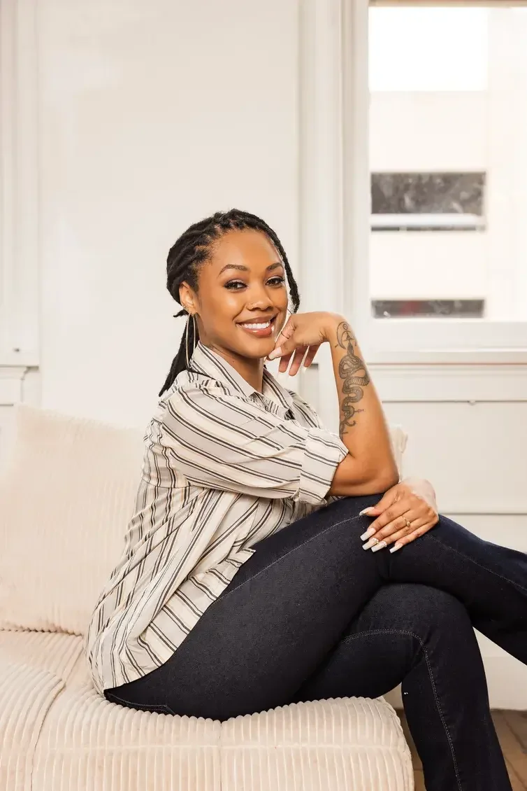 A woman with dreadlocks, tattoos, wearing a white dress, sitting on a black leather couch with decorative pillows, smiling and resting her chin on her hand. She is Skye Adams-Thomas, an LPC Associate in Garland Texas providing therapy to teens.