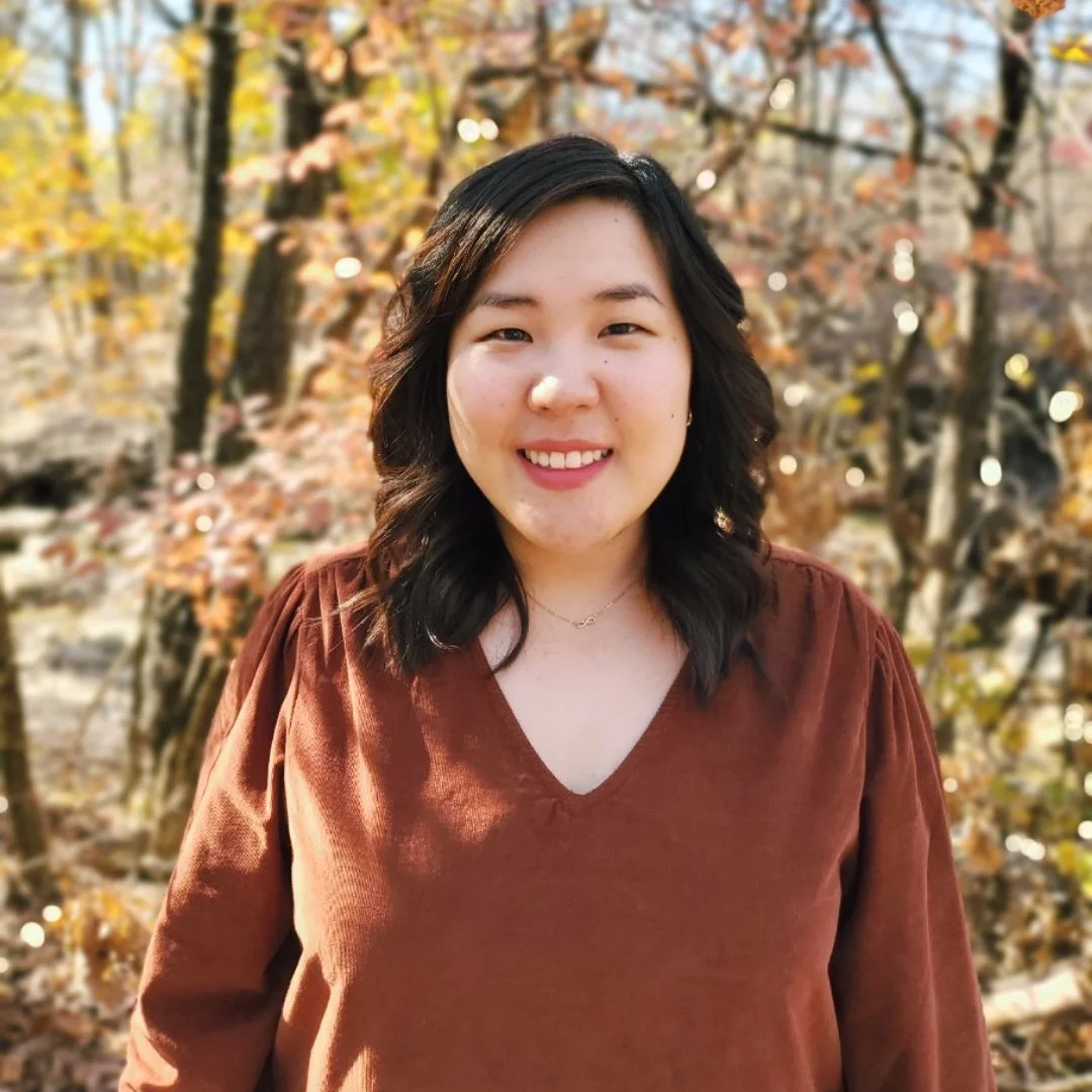 A young woman with shoulder-length dark hair smiling outdoors during autumn, wearing a brown V-neck top, with trees and fall foliage in the background.