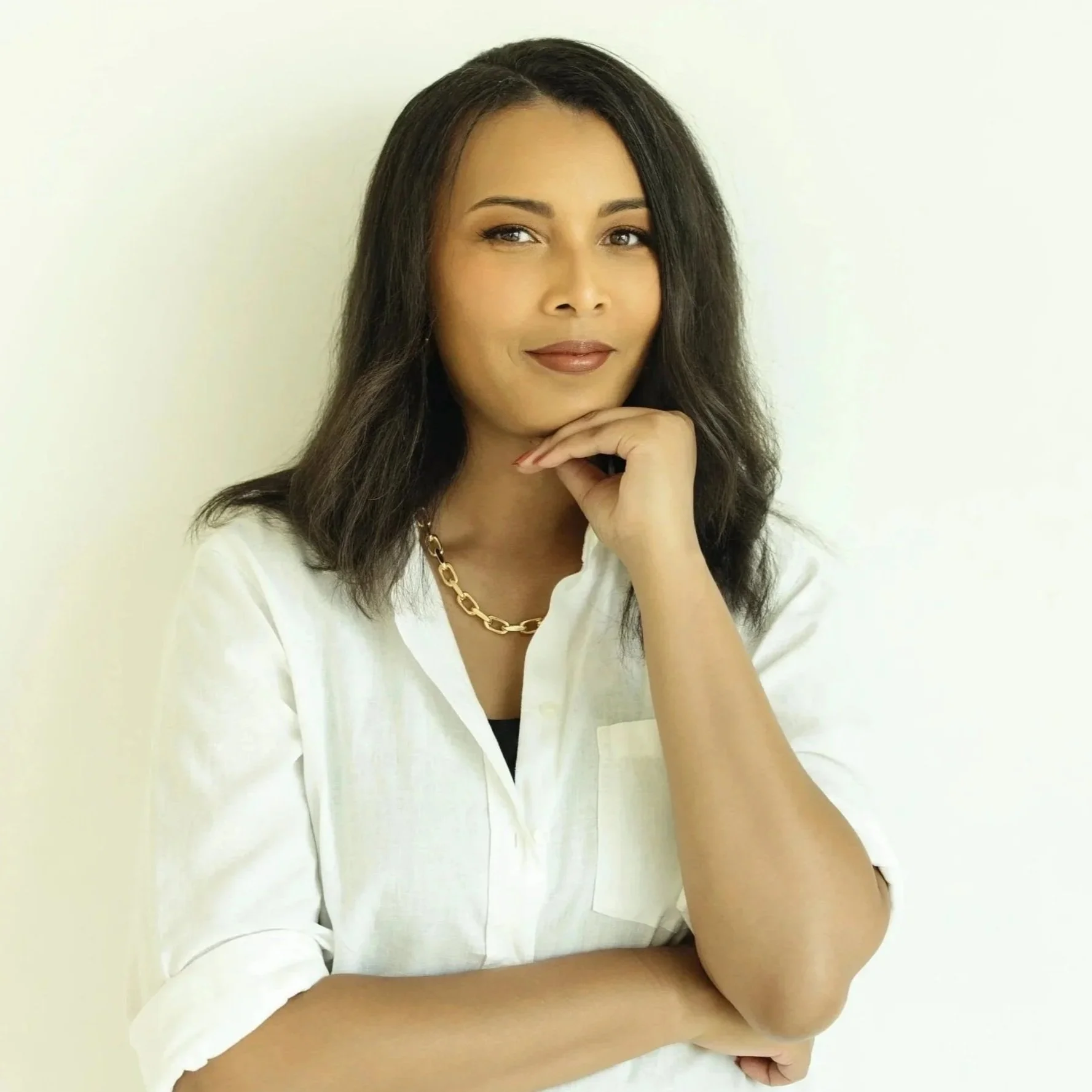 Portrait of a woman with shoulder-length dark hair, wearing a white shirt and gold chain necklace, posing with her chin resting on her hand against a plain light-colored background.