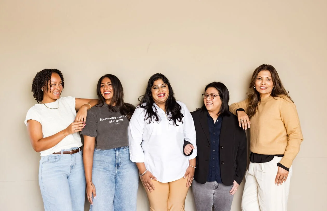Five women standing against a beige wall, smiling and casual, with arms around each other.