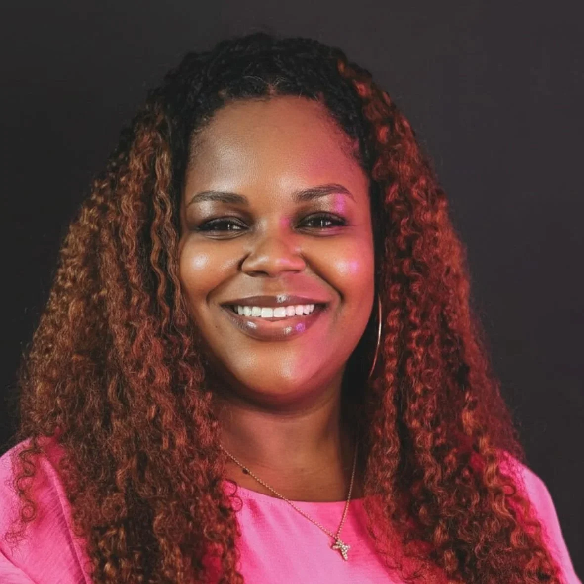 Smiling person with braided hair wearing a pink shirt outdoors with trees and buildings in the background.