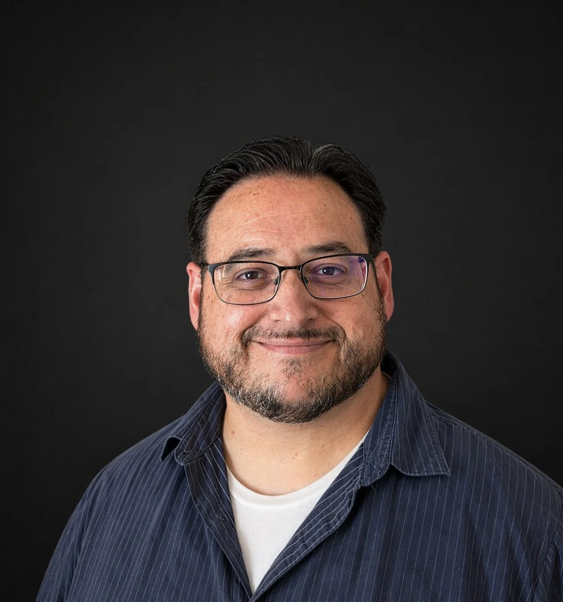 Headshot of a smiling man with glasses, dark hair, a beard, wearing a navy blue striped shirt against a black background.