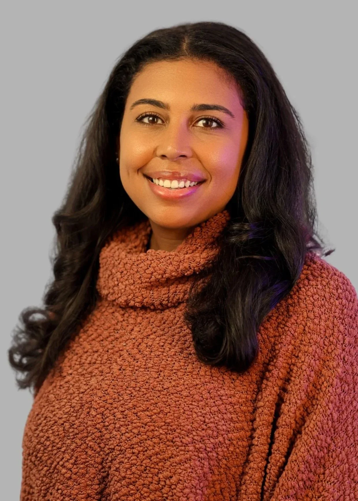 Portrait of a smiling woman with curly dark hair, wearing a textured brown turtleneck sweater, against a gray background.