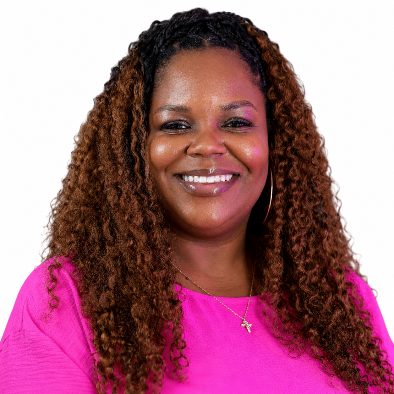 Smiling person with braided hair wearing a pink shirt outdoors with trees and buildings in the background.