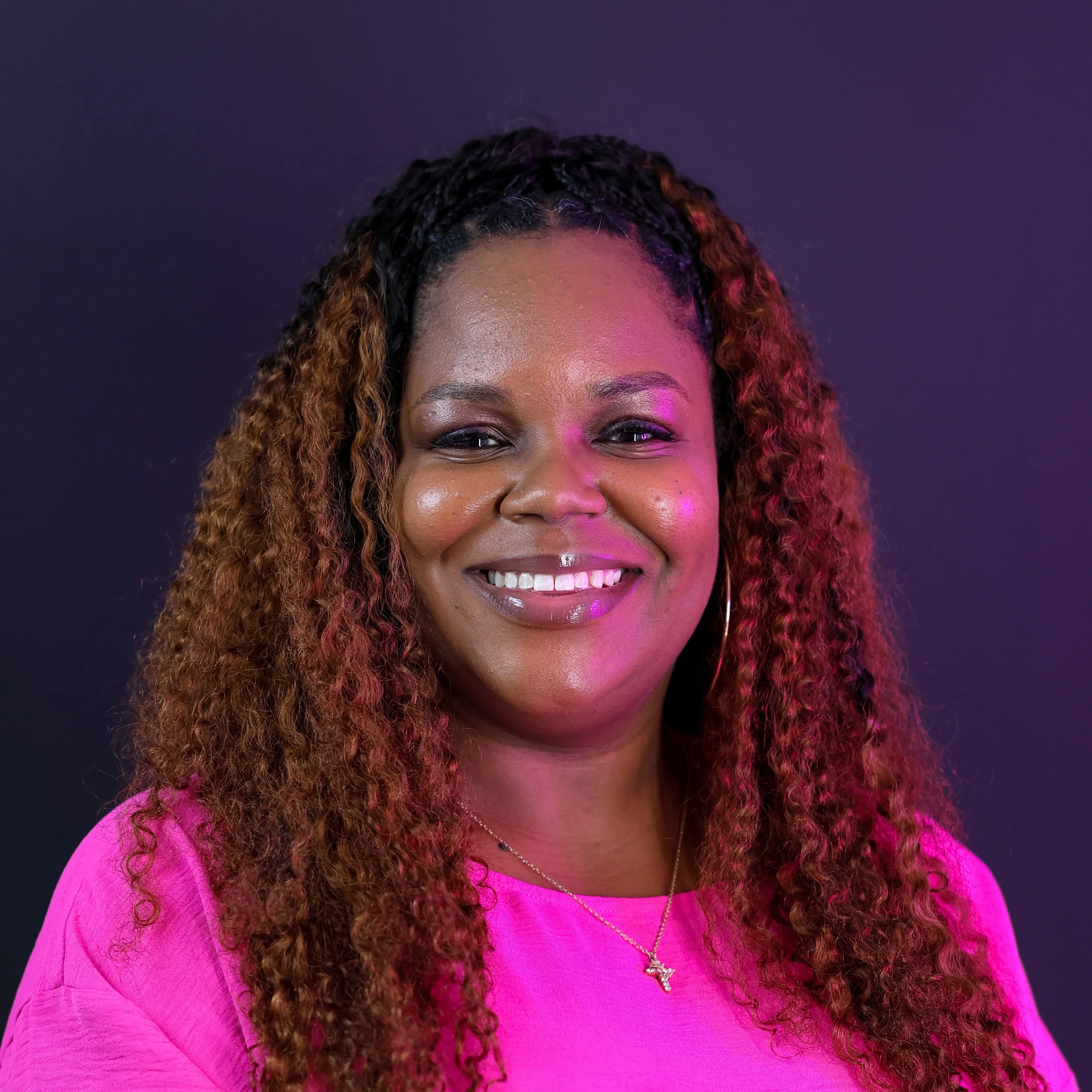 Smiling person with braided hair wearing a pink shirt outdoors with trees and buildings in the background.