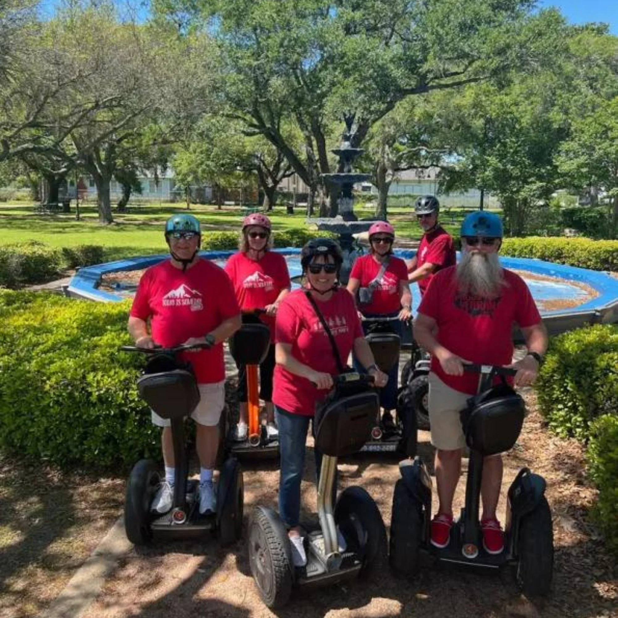 Group of people wearing helmets and red shirts standing with scooters in a park with trees, a fountain, and houses in the background.