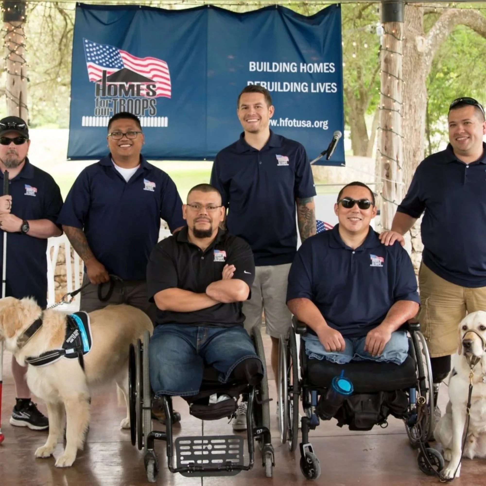Group of people, some in wheelchairs, with a large blue banner behind them that reads "Building Homes Rebuilding Lives" and features an American flag and a house graphic promoting Homes for Our Troops charity event.
