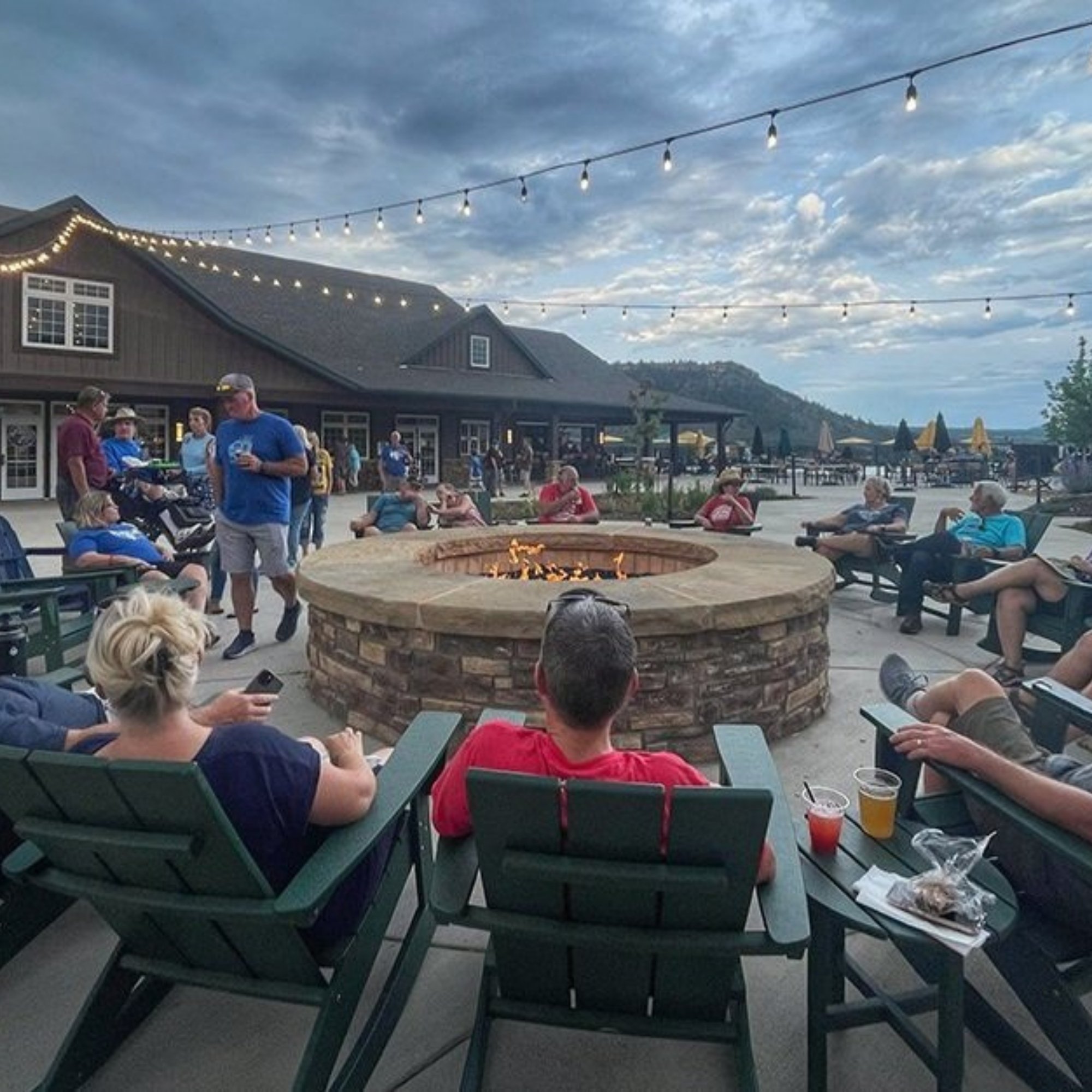 People gathered outdoors around a fire pit at dusk, with string lights overhead and a building with dark siding and windows in the background.